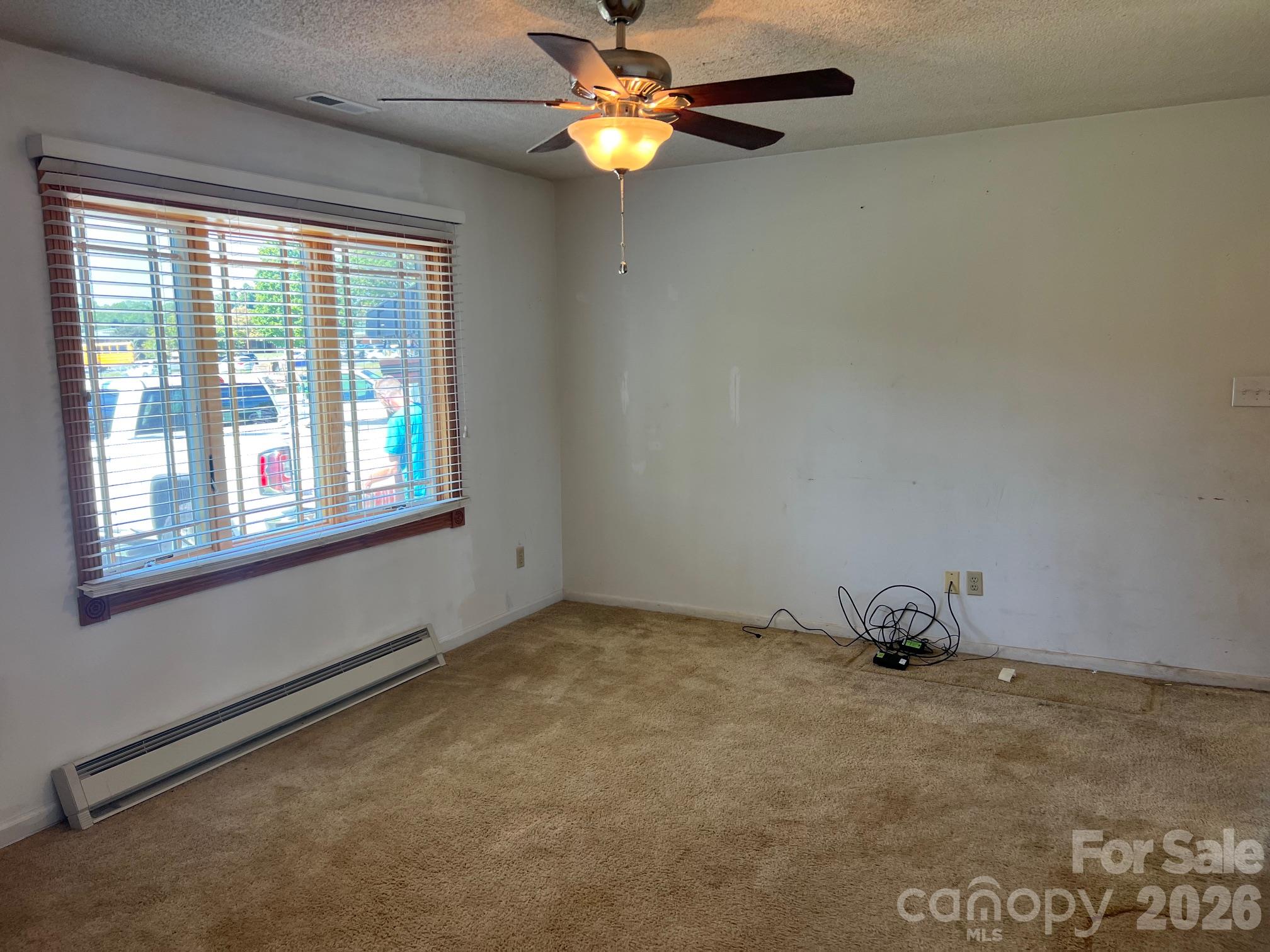 Unfurnished carpeted room with a large blinded window, ceiling fan, and electric baseboard heater.