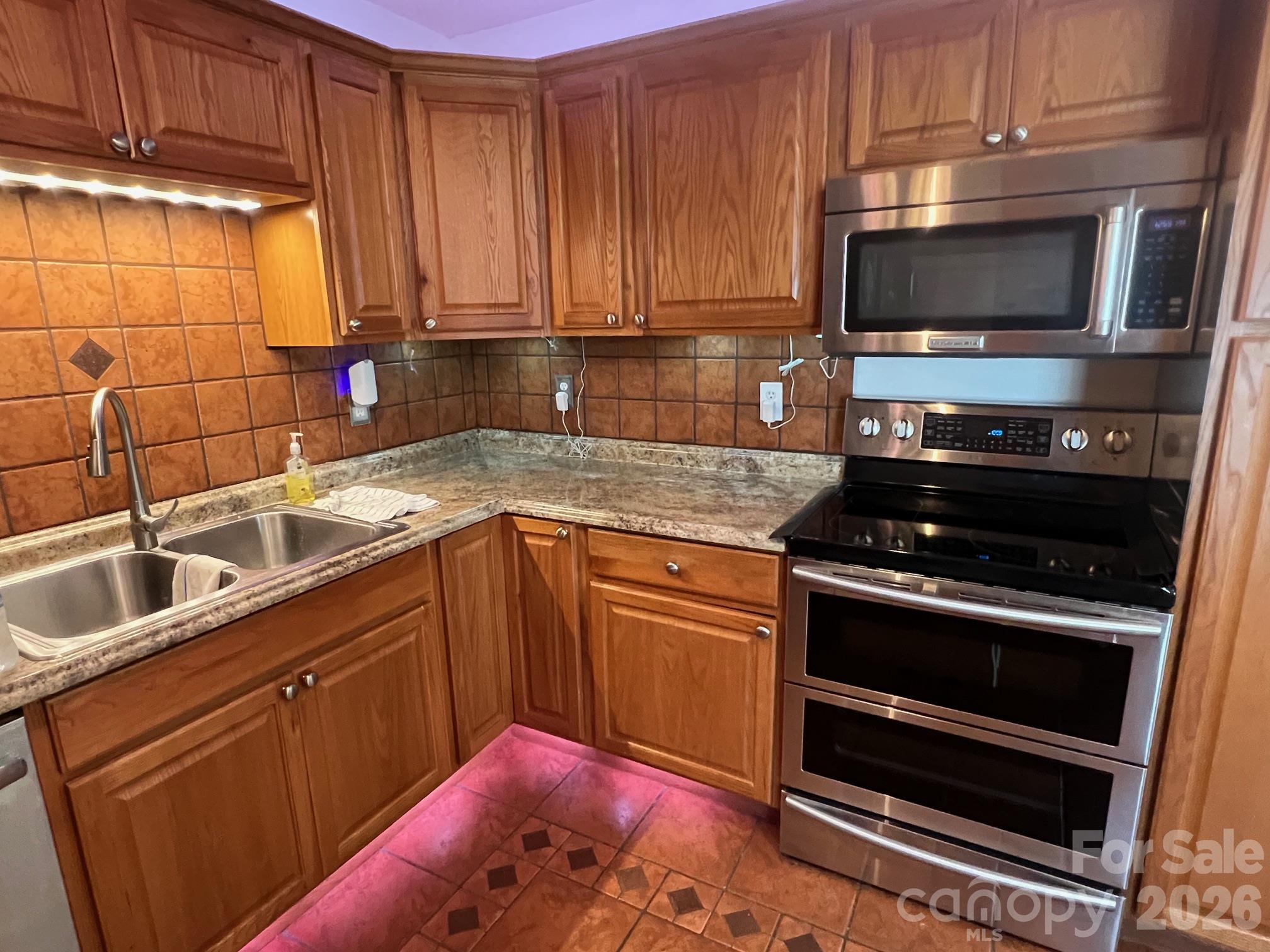Kitchen with warm wood cabinets, tiled backsplash, double sink, and stainless steel double oven.