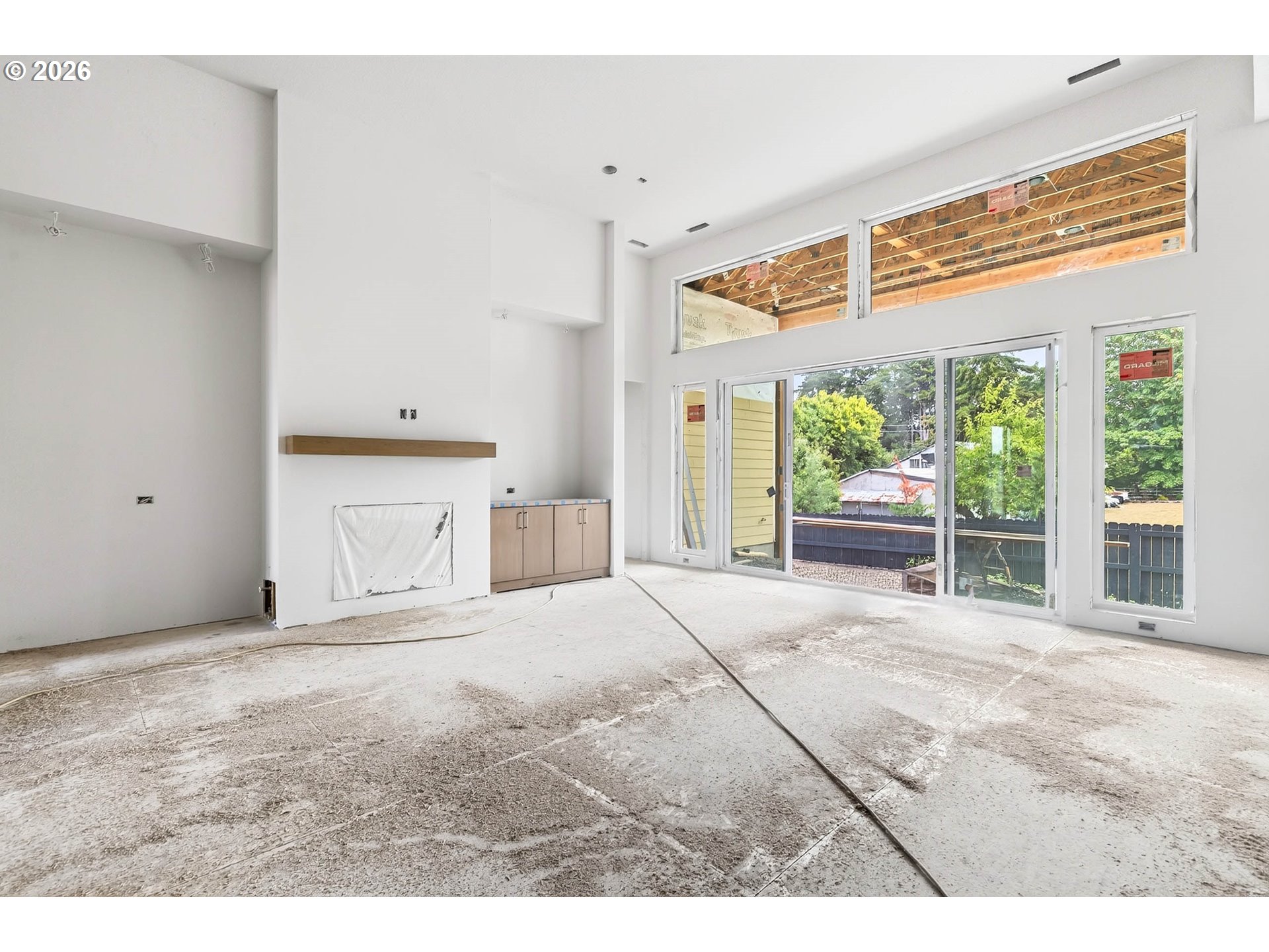 Unfinished living room with a white fireplace, exposed ceiling framing, and large windows overlooking a fenced yard.