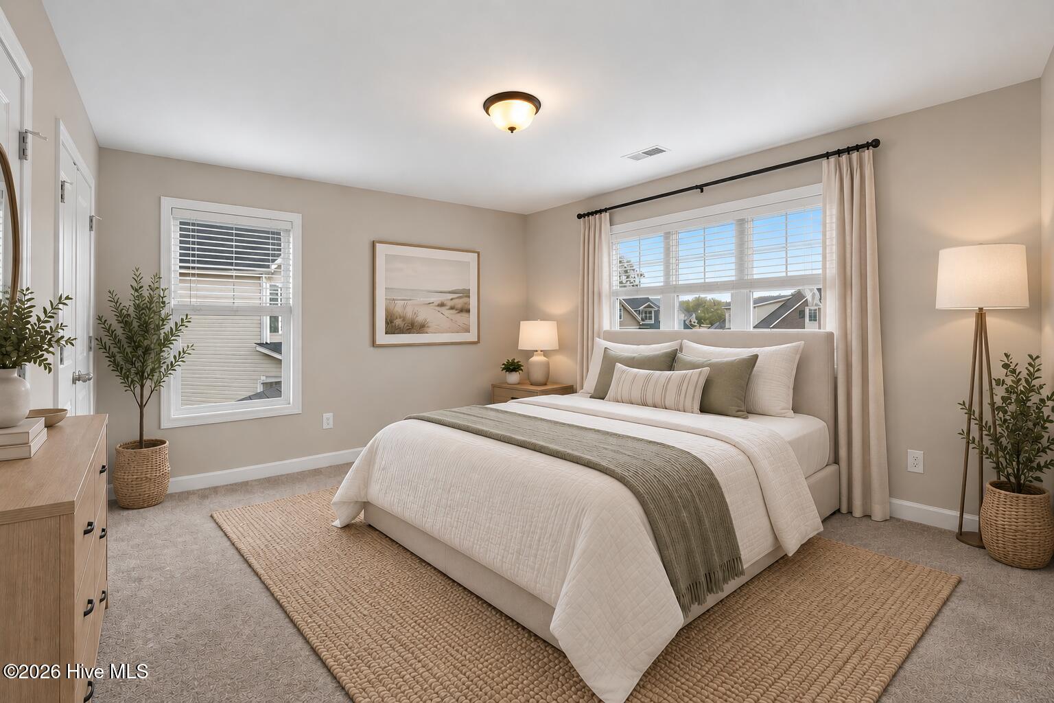 Staged bedroom with a white bed, muted olive throw, woven rug, and wide curtained windows.