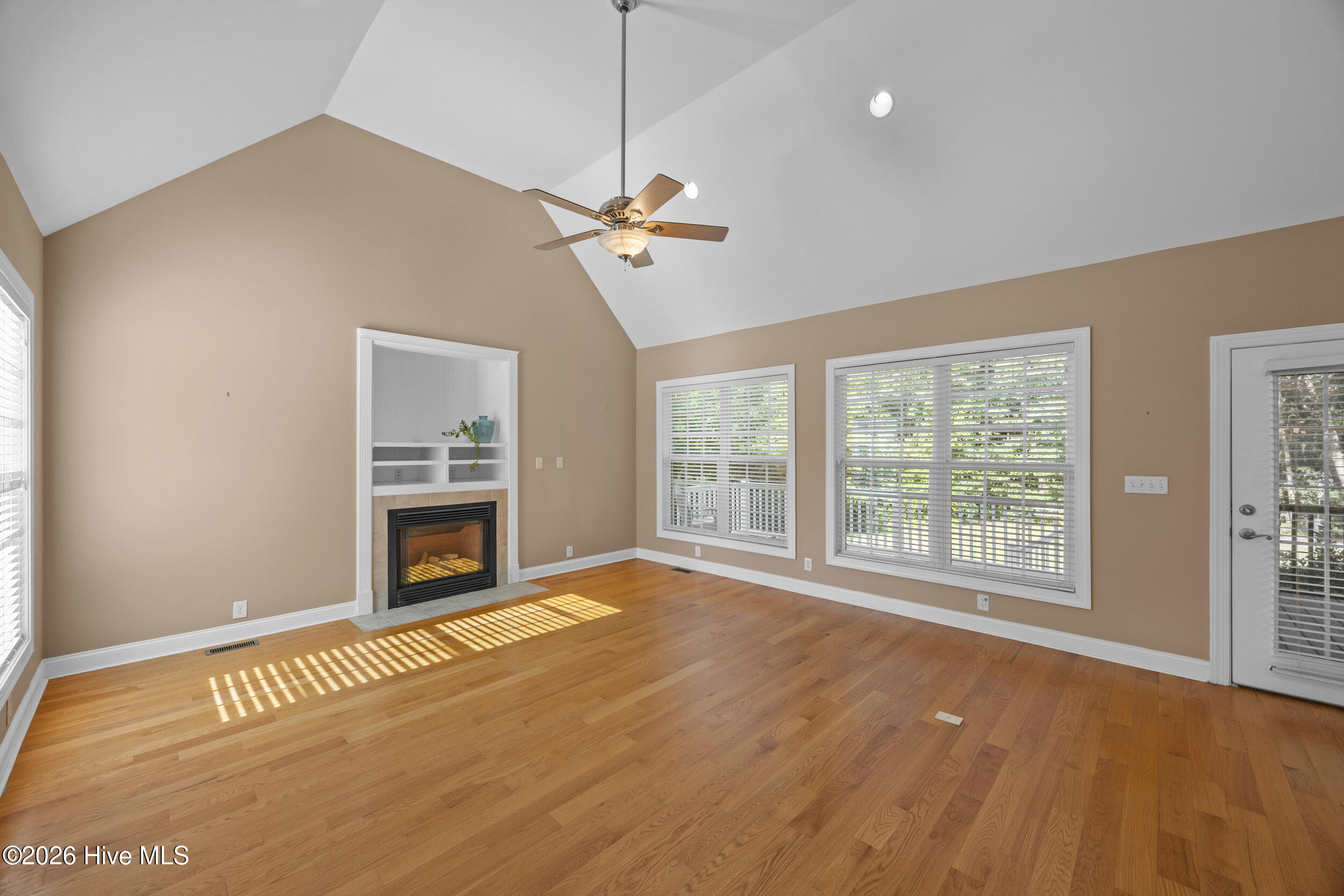 Empty living room with vaulted ceiling, hardwood floors, fireplace, and large windows with blinds.