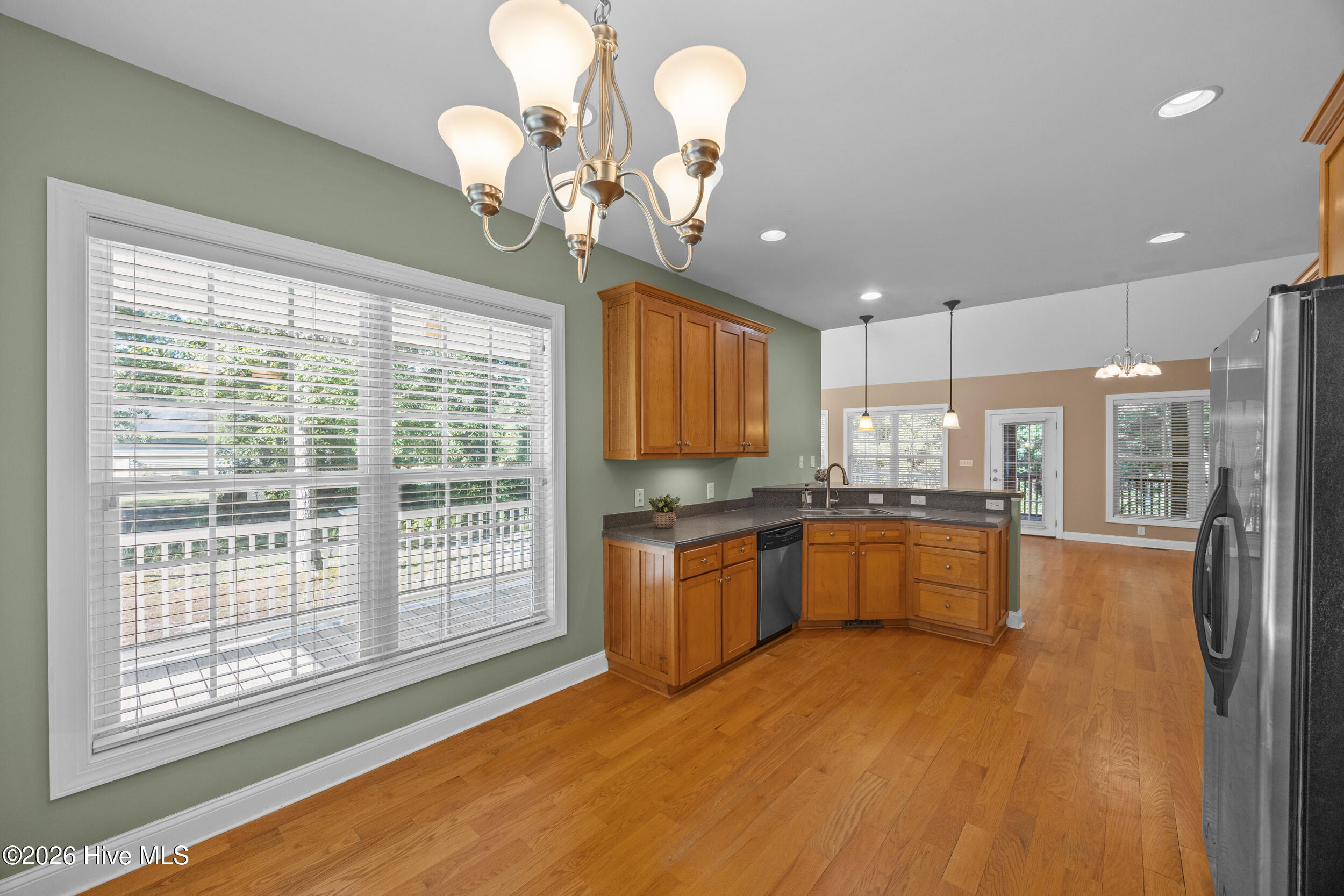 Kitchen with honey-toned cabinets, dark countertops, stainless steel refrigerator, and hardwood floors opening to a dining area.