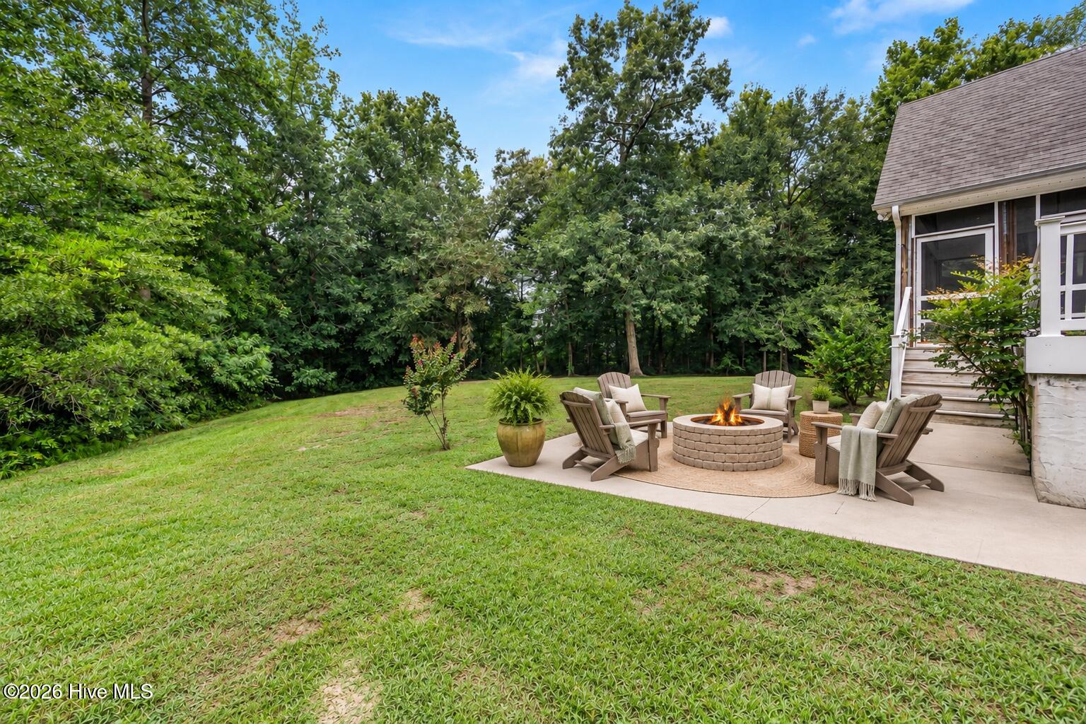 Backyard patio with cushioned chairs around a stone fire pit beside a broad lawn and wooded tree line.