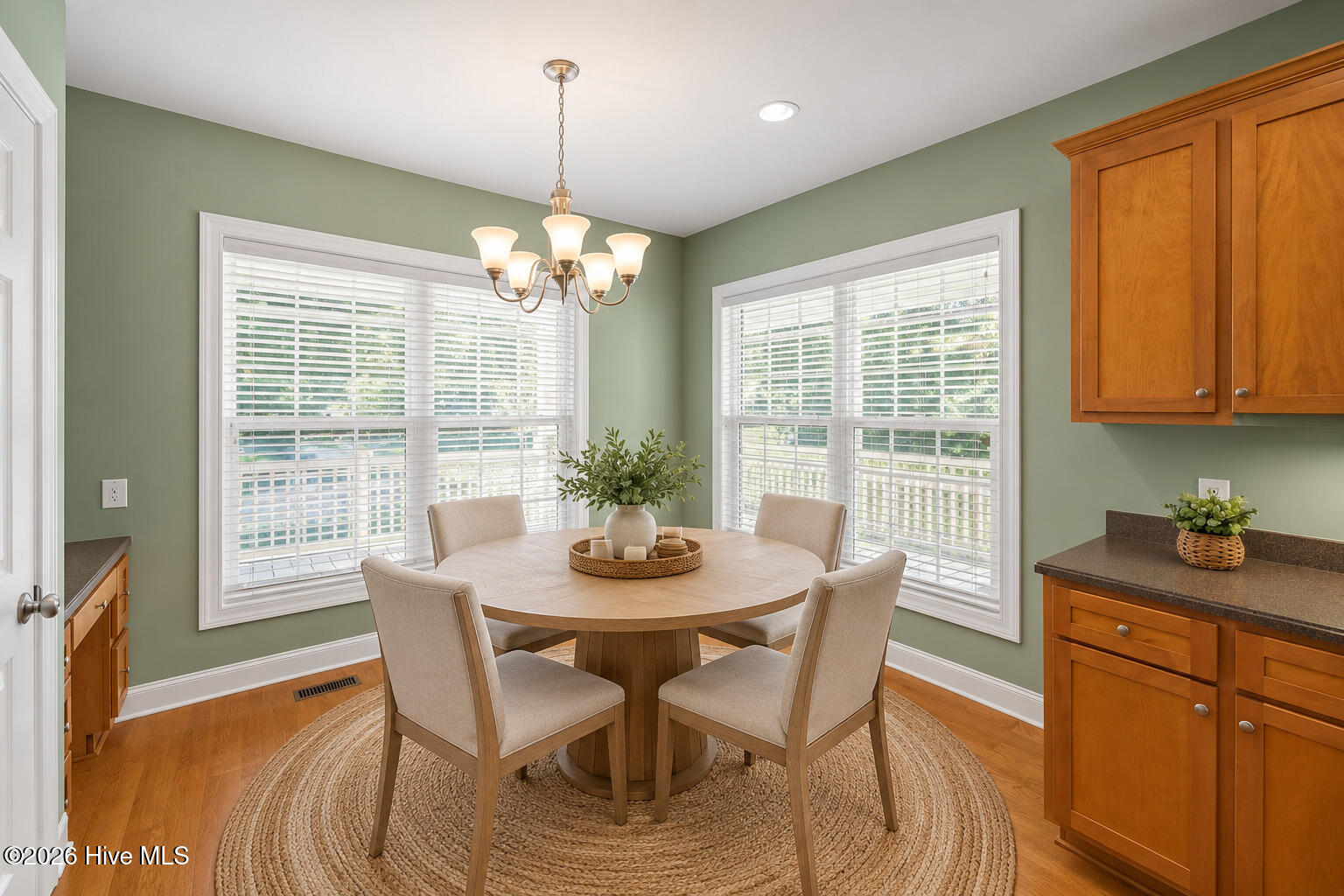 Dining area with a round table beneath a chandelier, sage green walls, large windows, and wood cabinetry.