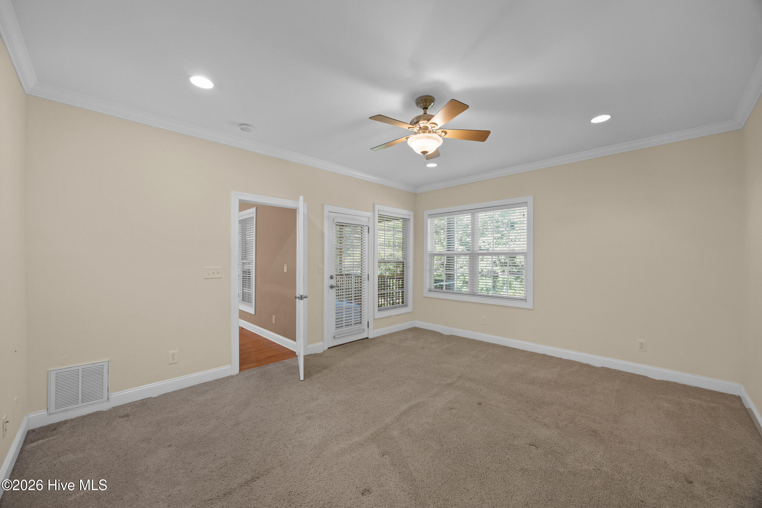 Empty carpeted room with a ceiling fan, shuttered glass door, and wide windows with blinds.