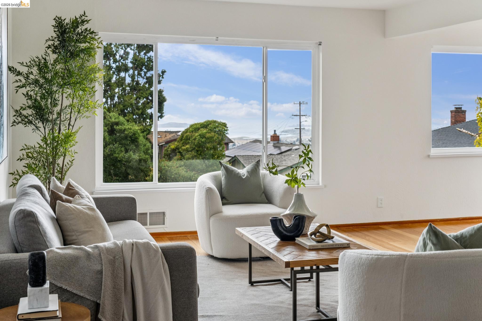 Staged living room with a large picture window overlooking rooftops, trees, and distant water.
