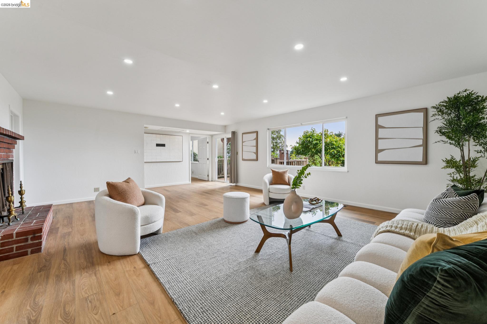 Staged living room with a brick fireplace, wood floors, and large windows overlooking leafy trees.