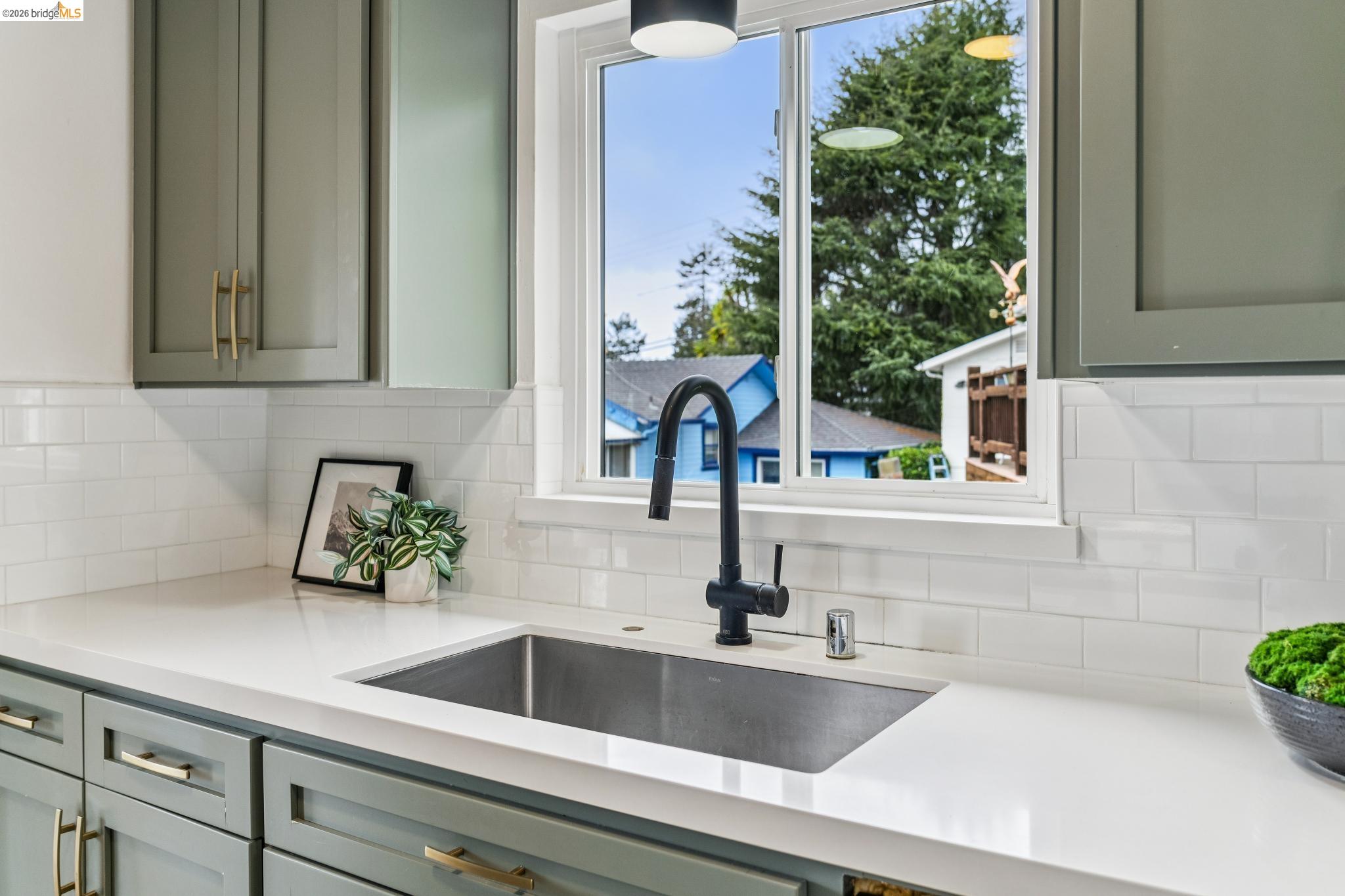 Kitchen sink beneath a large window, with sage-green cabinets, brass hardware, and white subway tile.