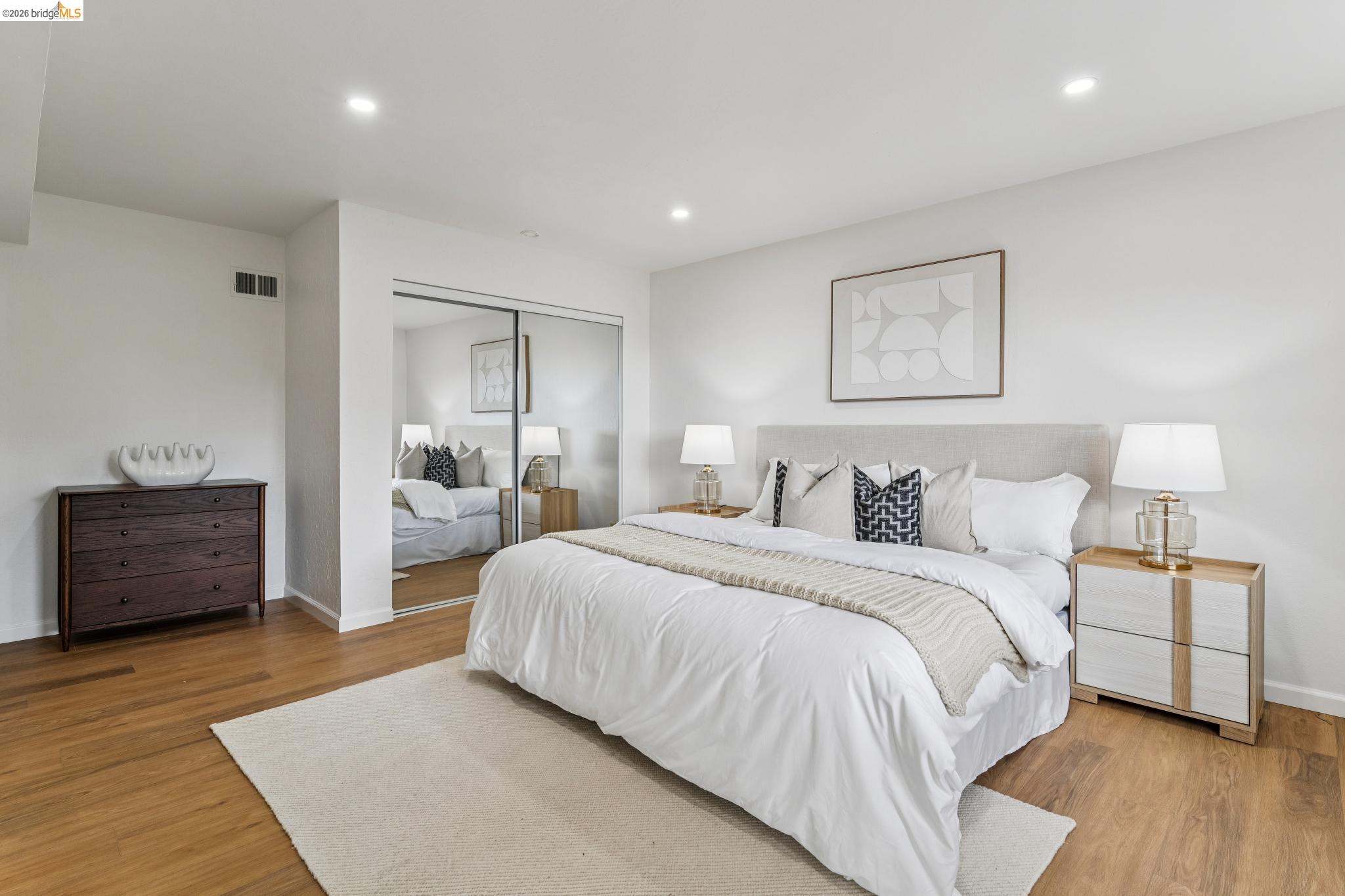 Staged bedroom with a white bed, mirrored closet doors, and warm hardwood floors.
