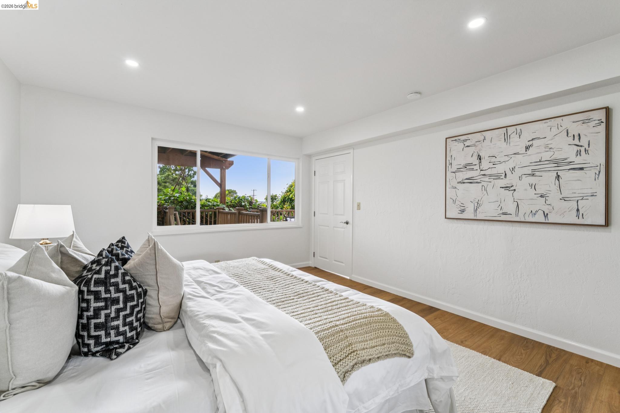 Staged bedroom with a white bed and a large window overlooking a leafy deck.