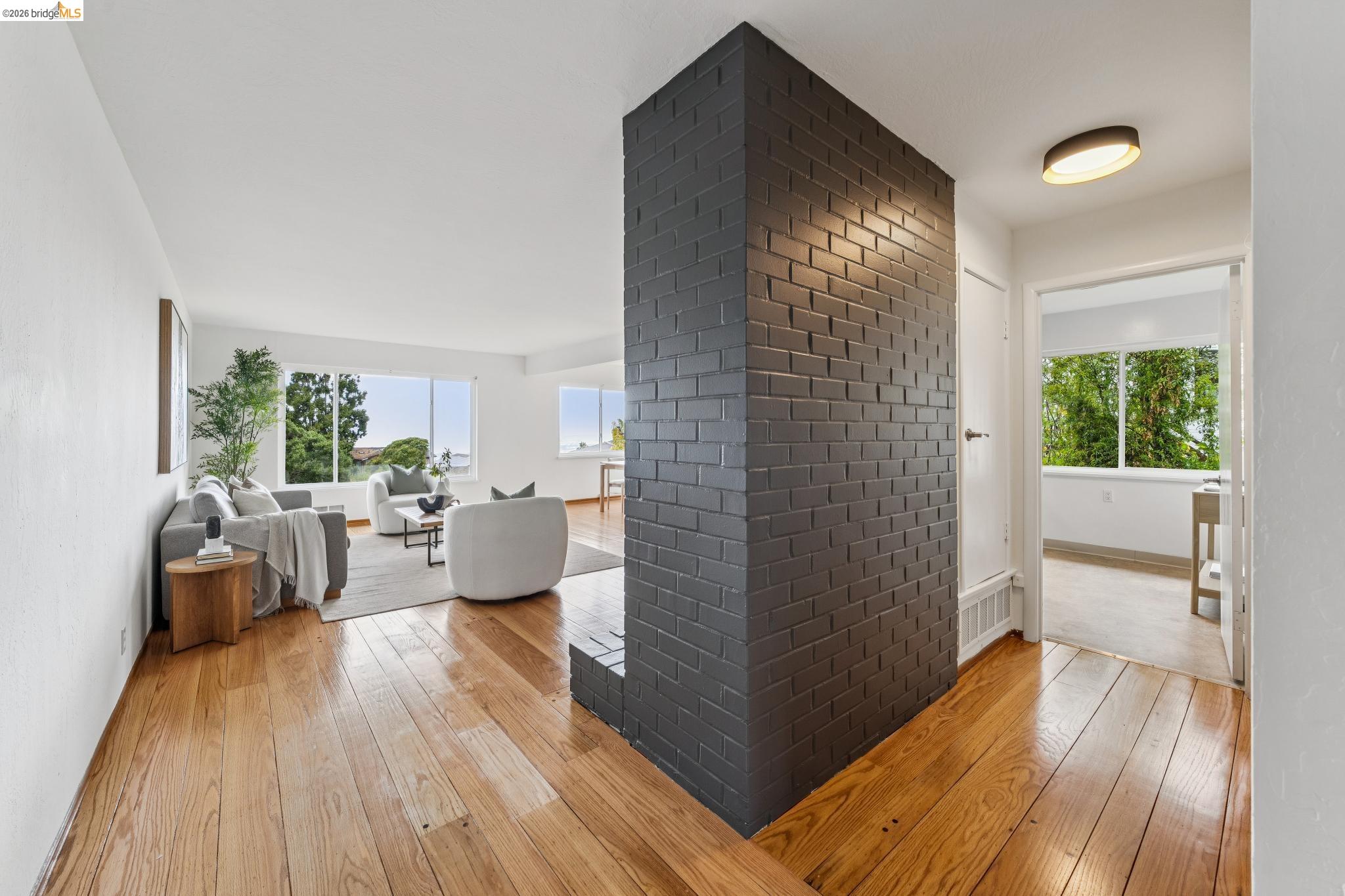 Staged living room with hardwood floors, large windows, and a prominent dark brick chimney.