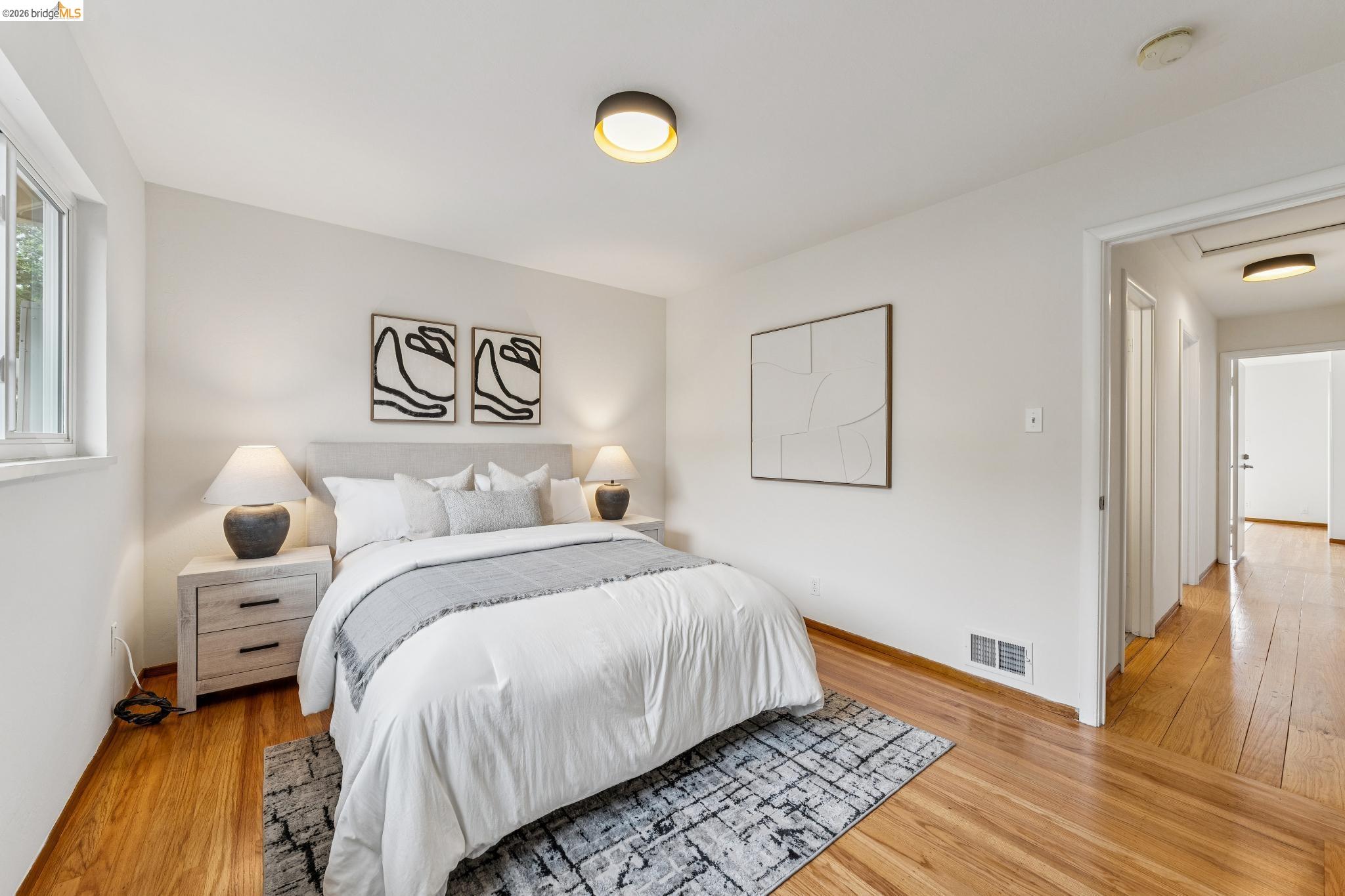 Staged bedroom with a white bed, gray patterned rug, hardwood floors, and an open doorway to the hall.