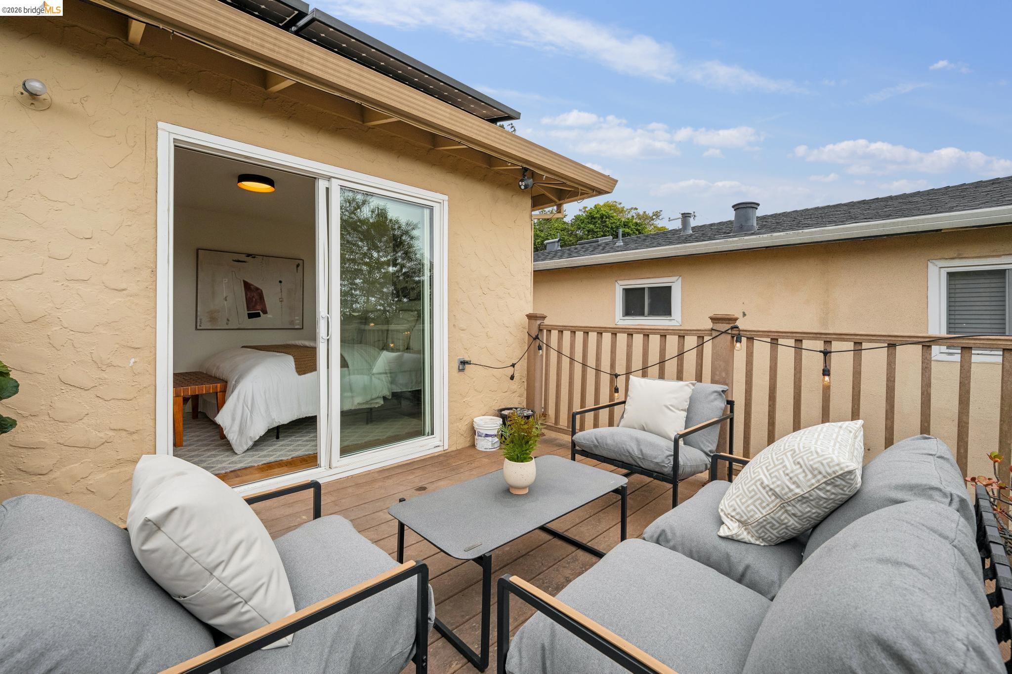 Furnished wooden deck with gray lounge seating, string lights, and an open sliding door to a bedroom.