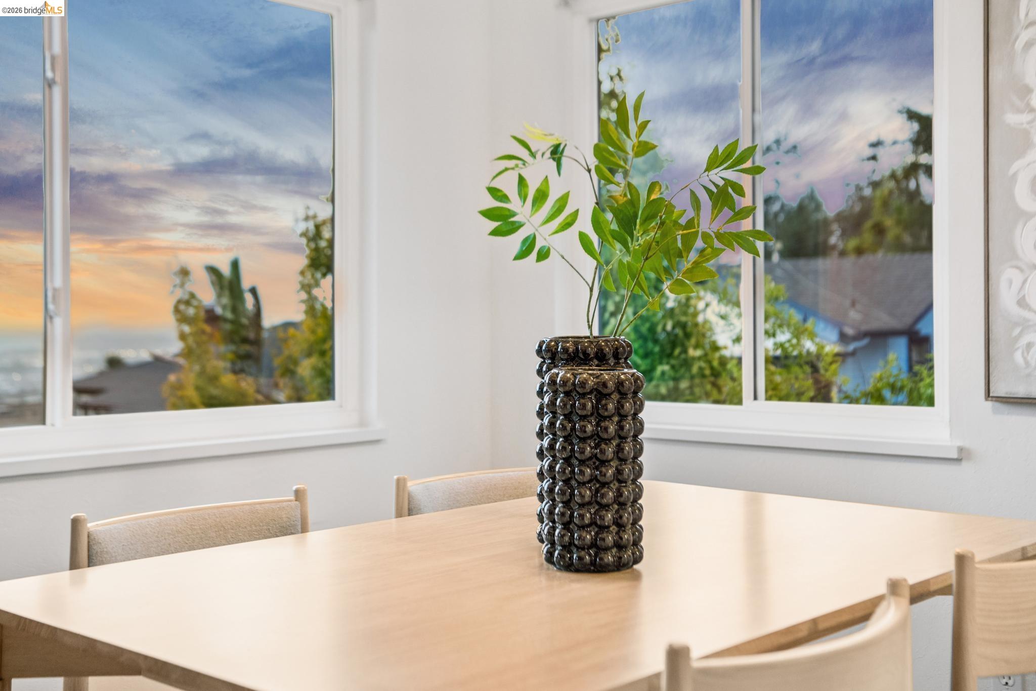 Dining area with a light wood table, sculptural vase, and large windows overlooking water.