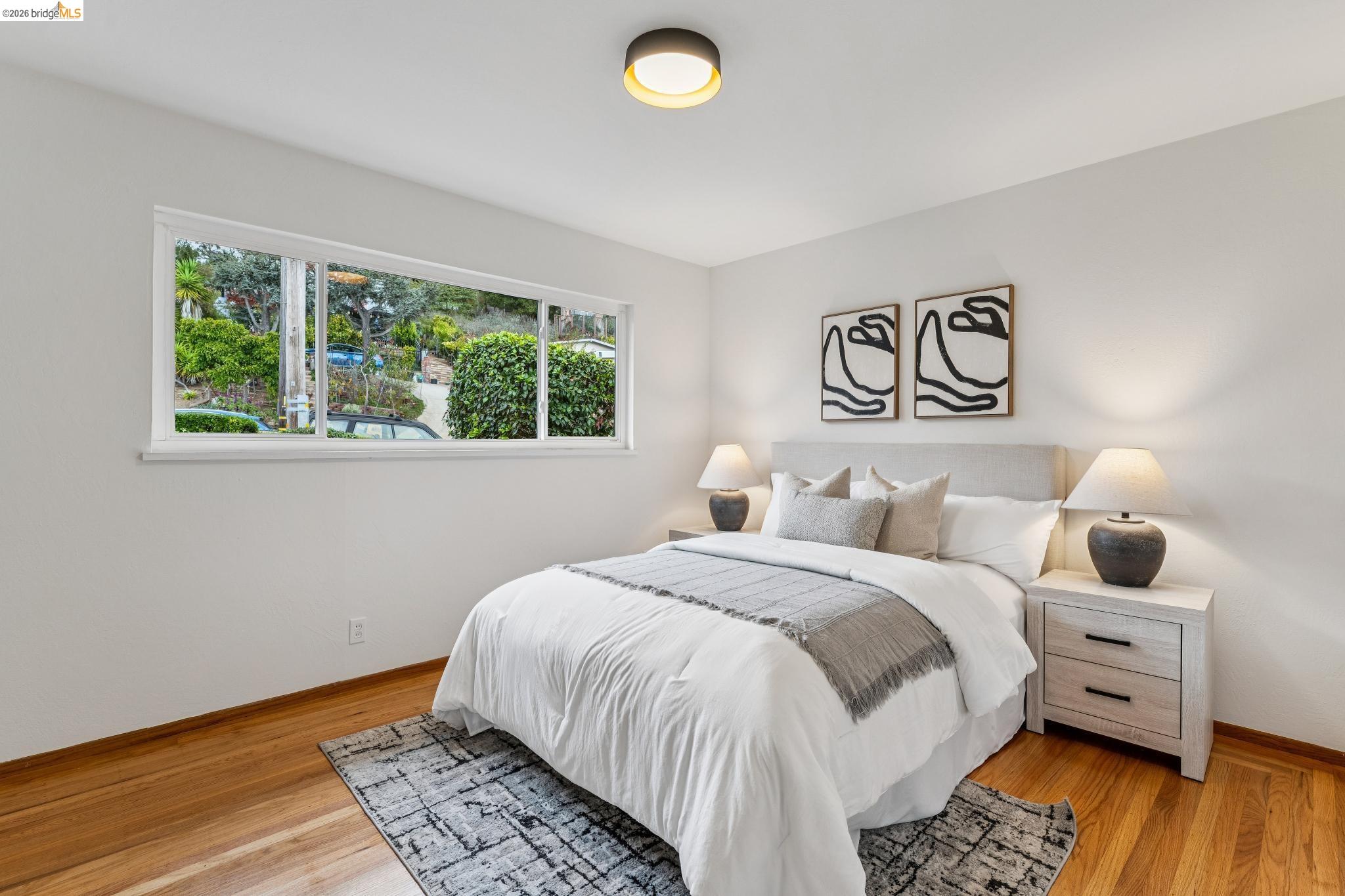 Staged bedroom with a white bed, matching lamps, hardwood floors, and a wide window overlooking greenery.