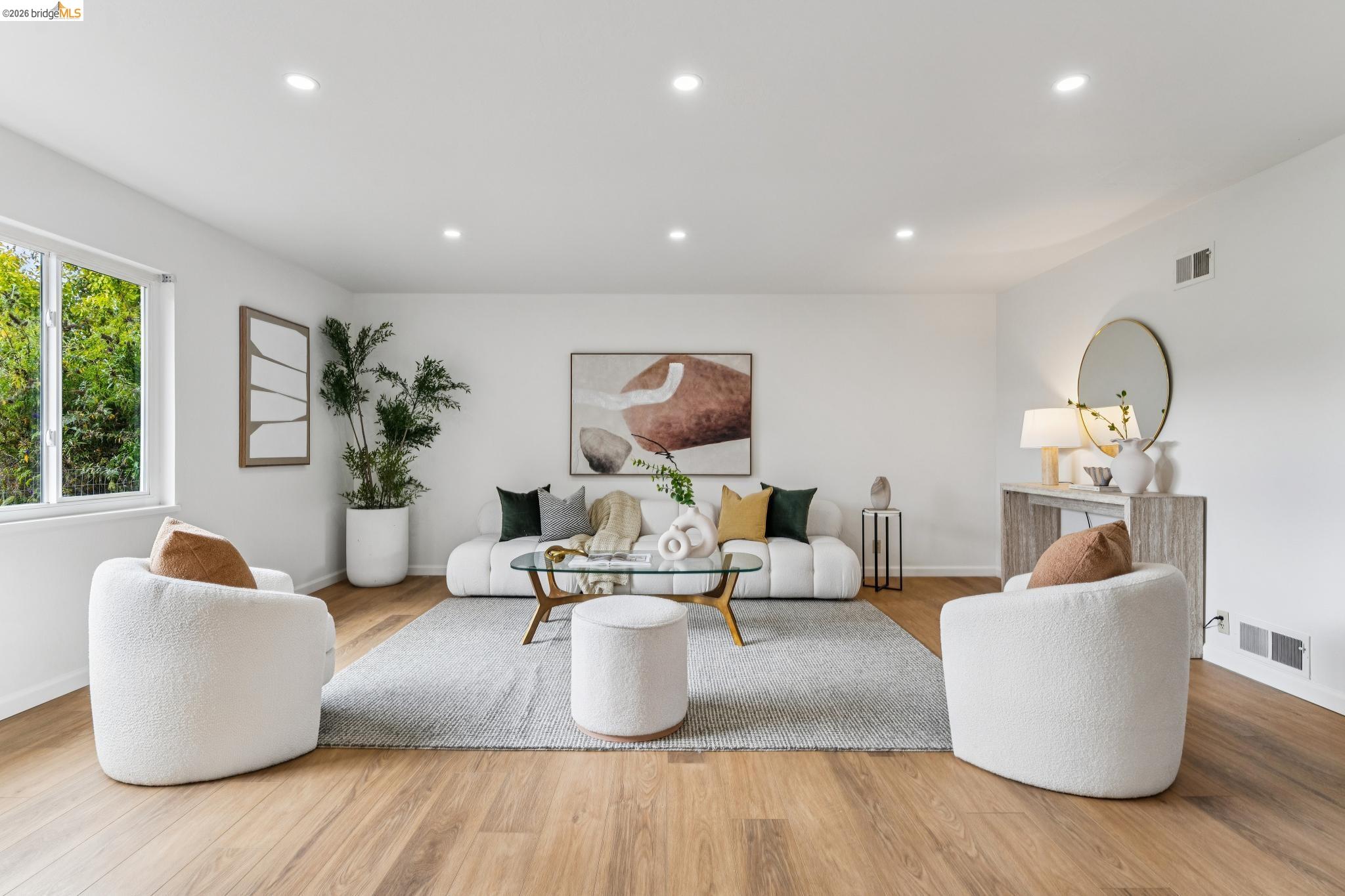 Staged living room with white seating, a glass coffee table, and a large window overlooking greenery.