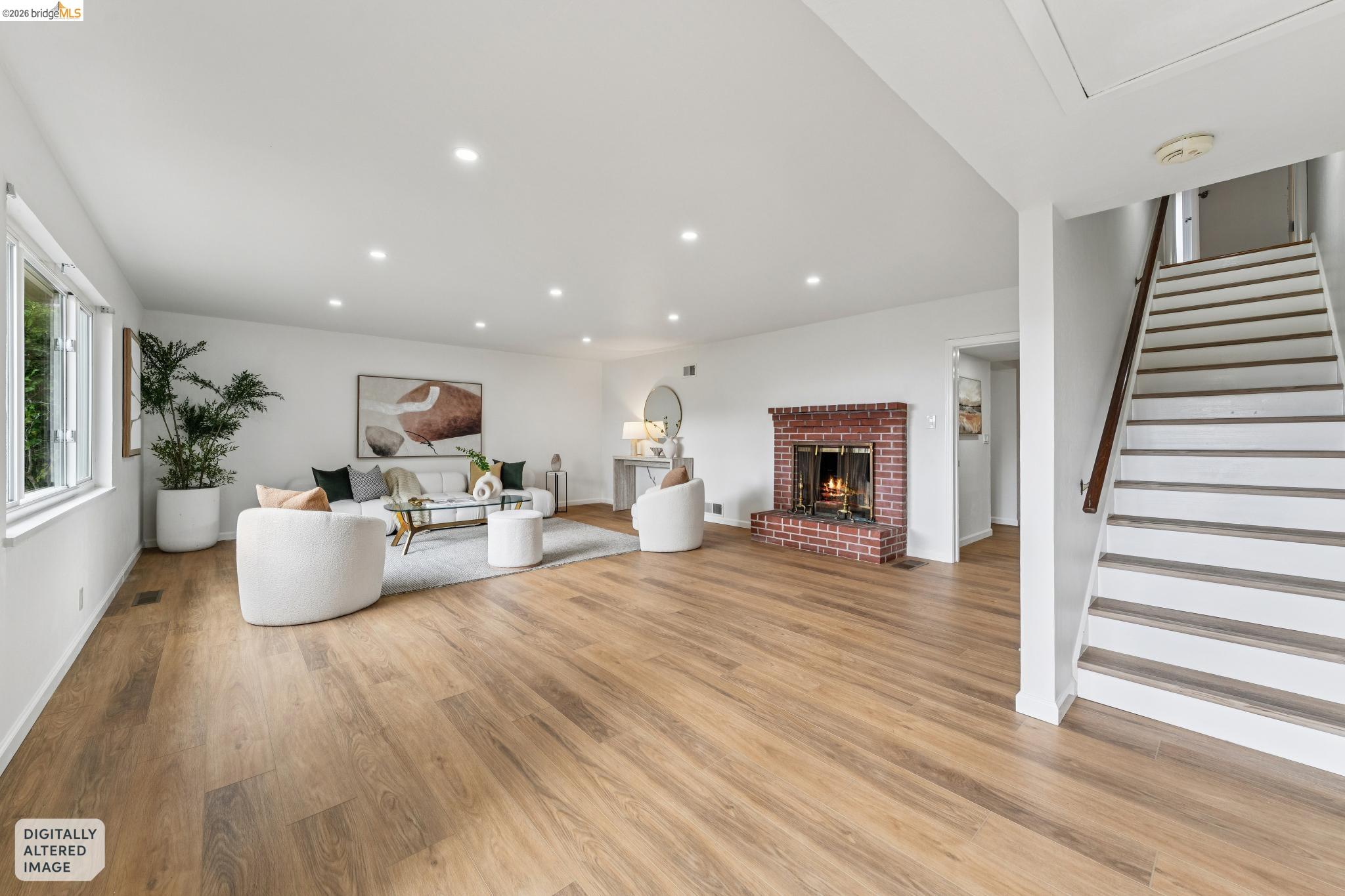 Staged living room with a brick fireplace, white seating, wood floors, and an open staircase.