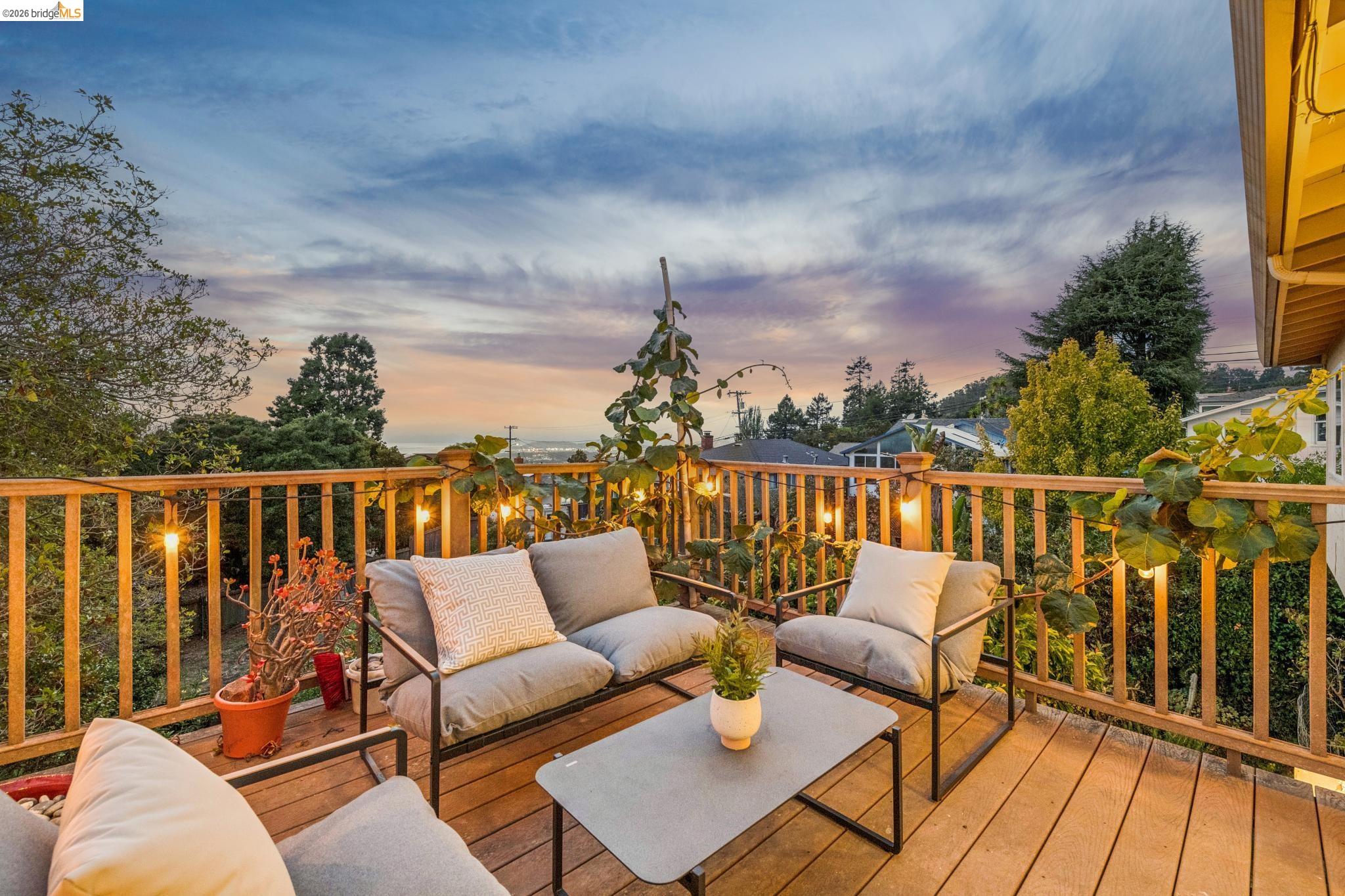 Furnished wooden deck with cushioned seating and string lights, overlooking trees and a distant water view.