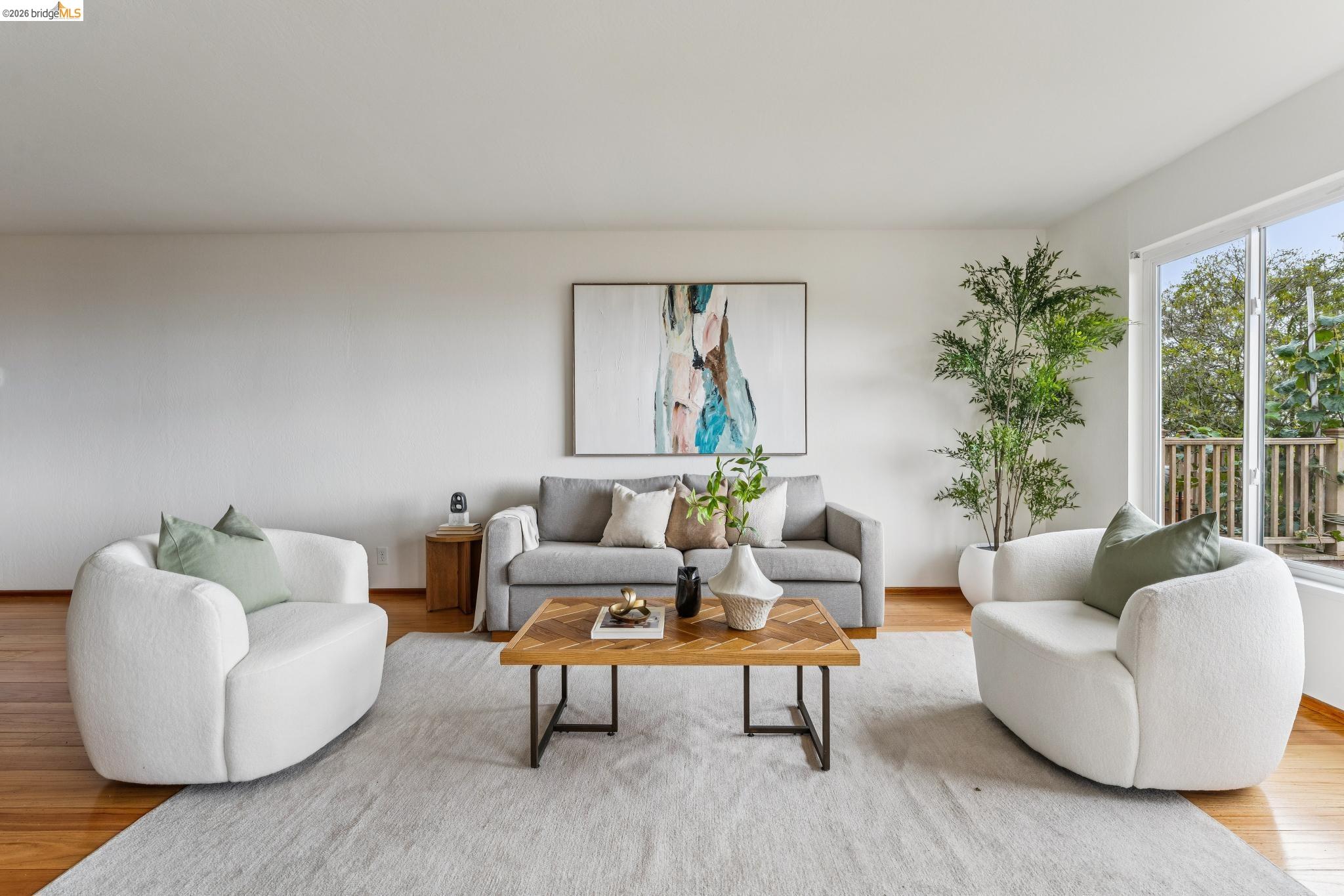 Staged living room with a gray sofa, white armchairs, and large windows overlooking a deck.