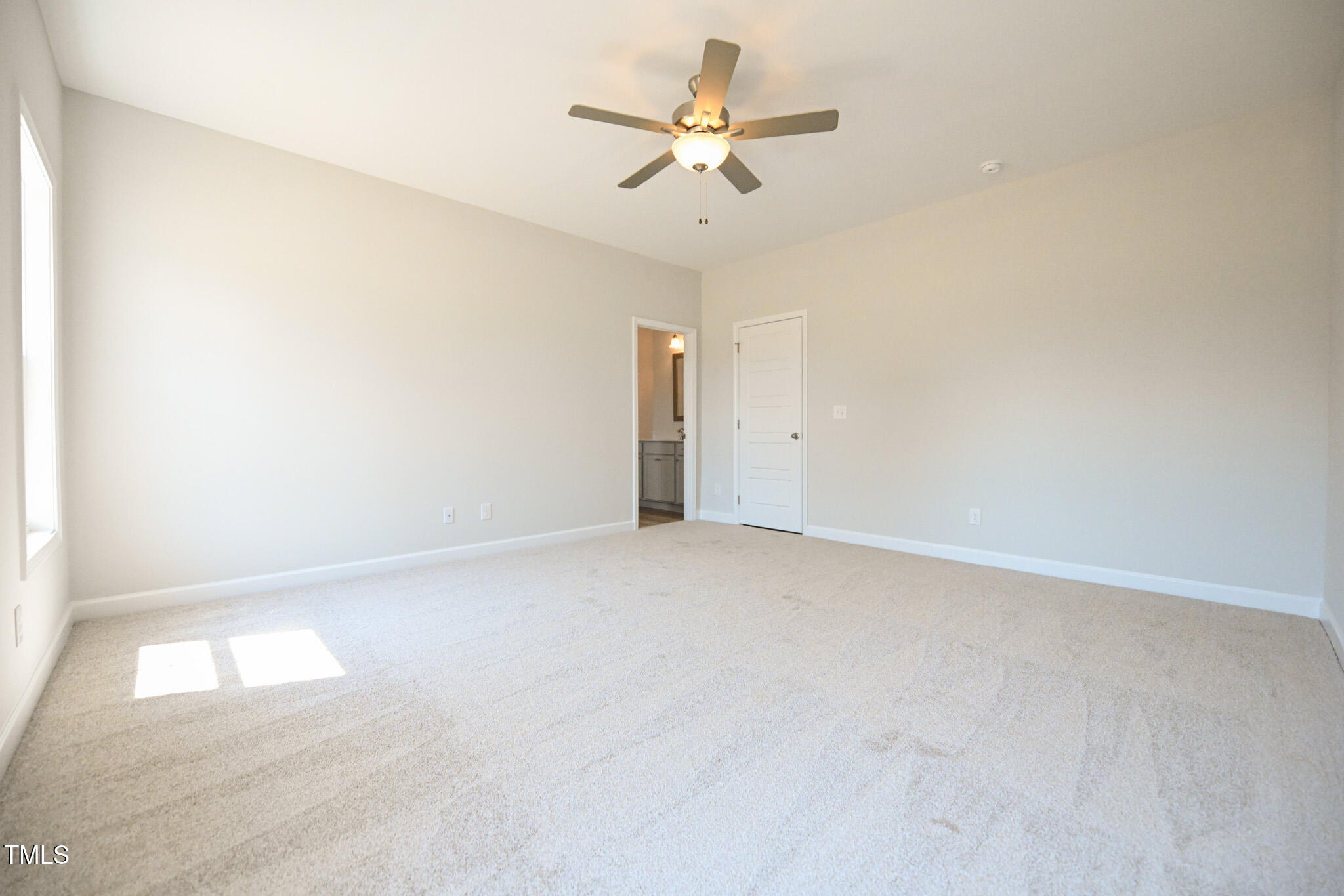 Empty carpeted bedroom with a ceiling fan and sunlight falling through the window.
