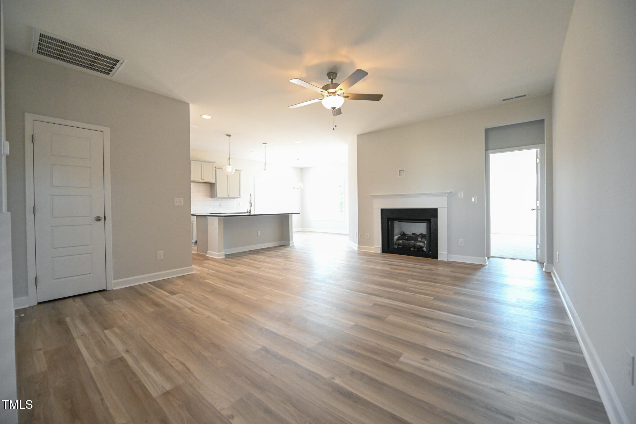 Empty living room with wood-look floors, a black fireplace, ceiling fan, and open kitchen beyond.