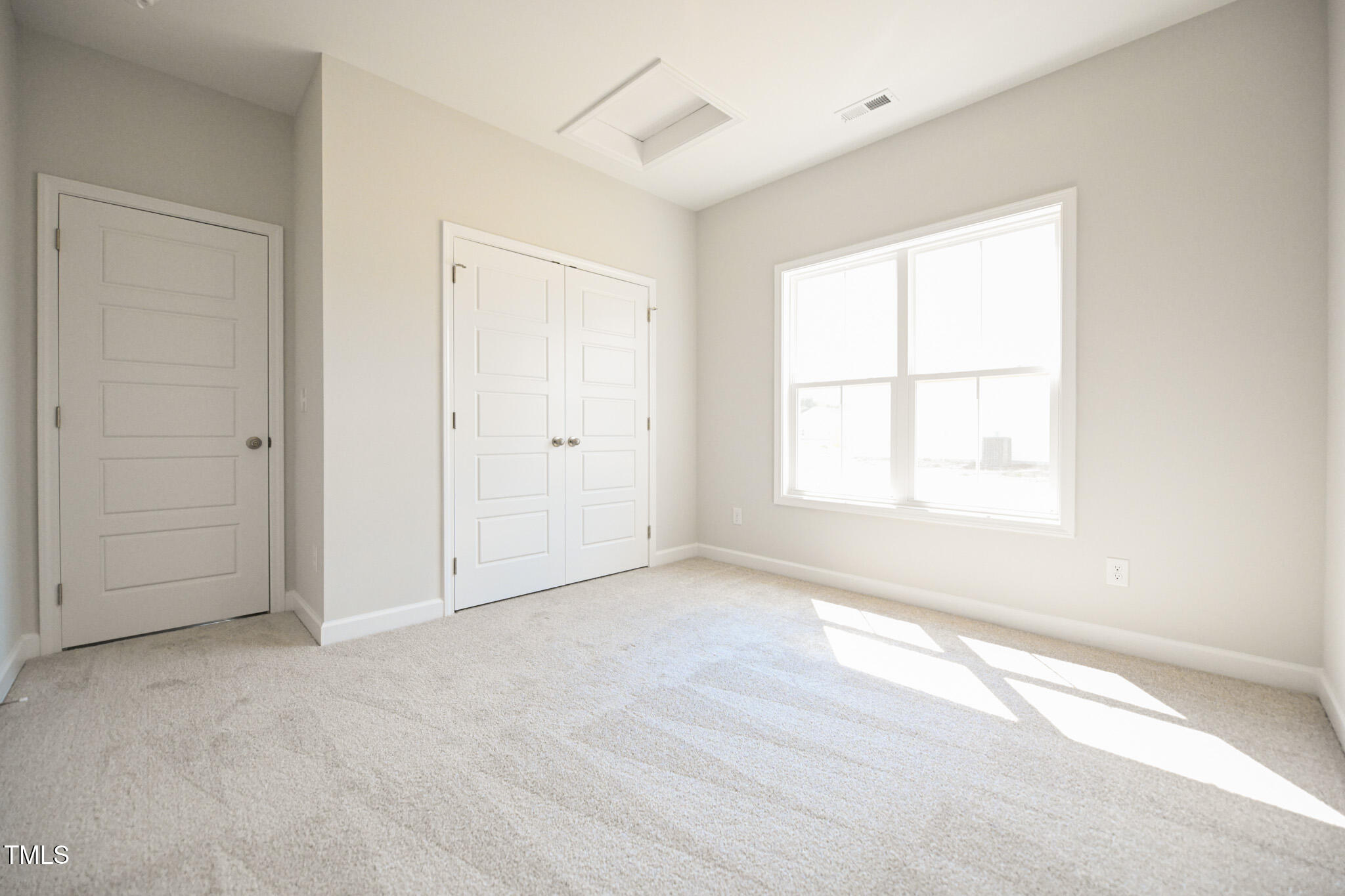 Empty carpeted bedroom with paneled closet doors and a large sunlit window.