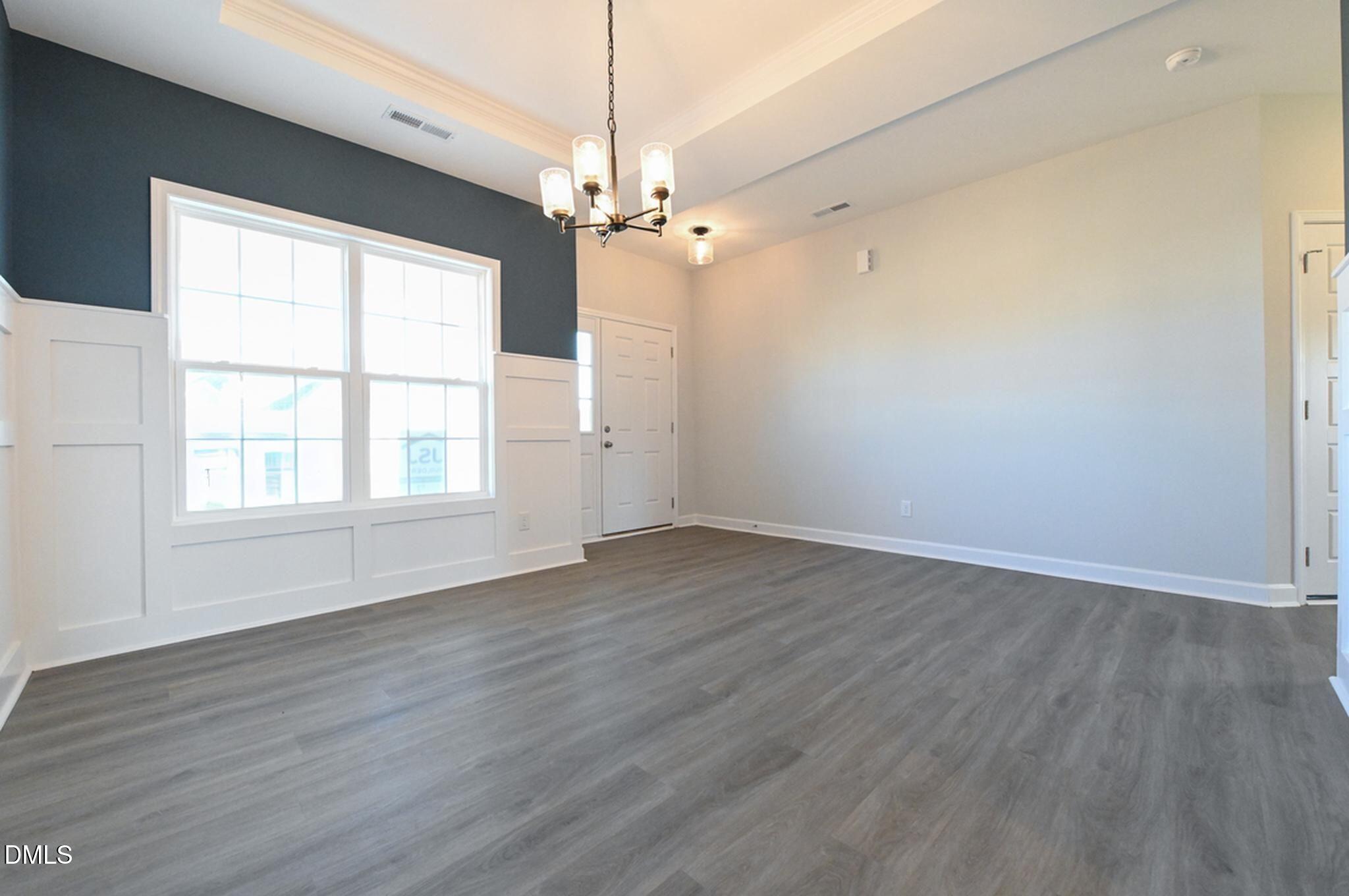 Empty living room with gray plank flooring, paneled walls, large windows, and a chandelier.