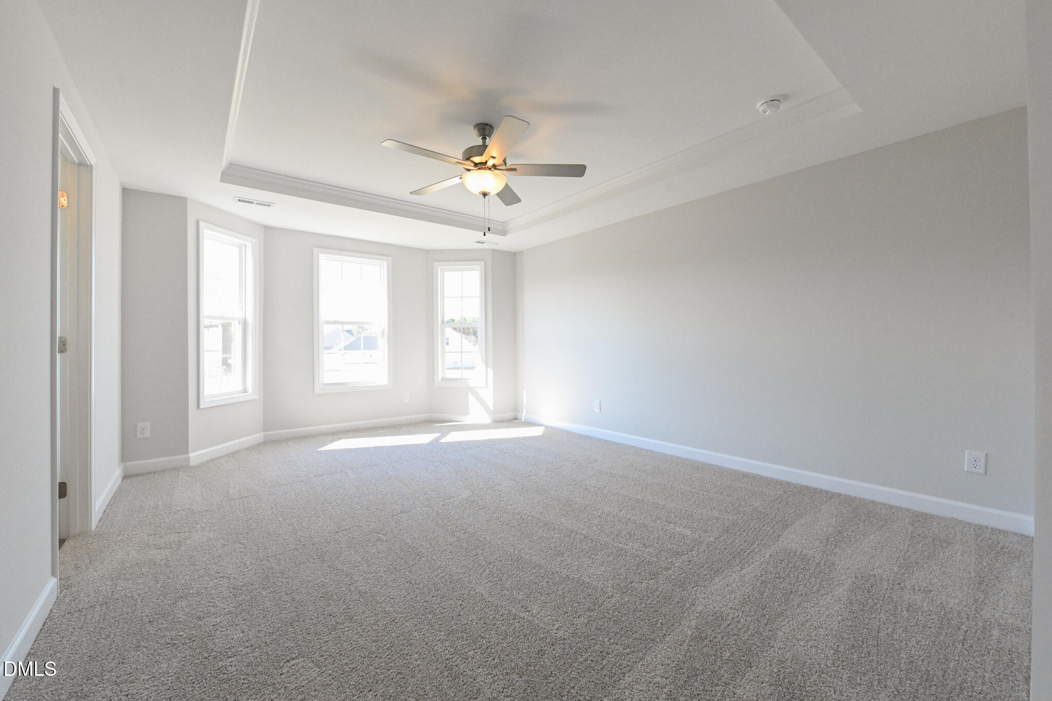 Empty carpeted room with three windows and a lit ceiling fan beneath a tray ceiling.