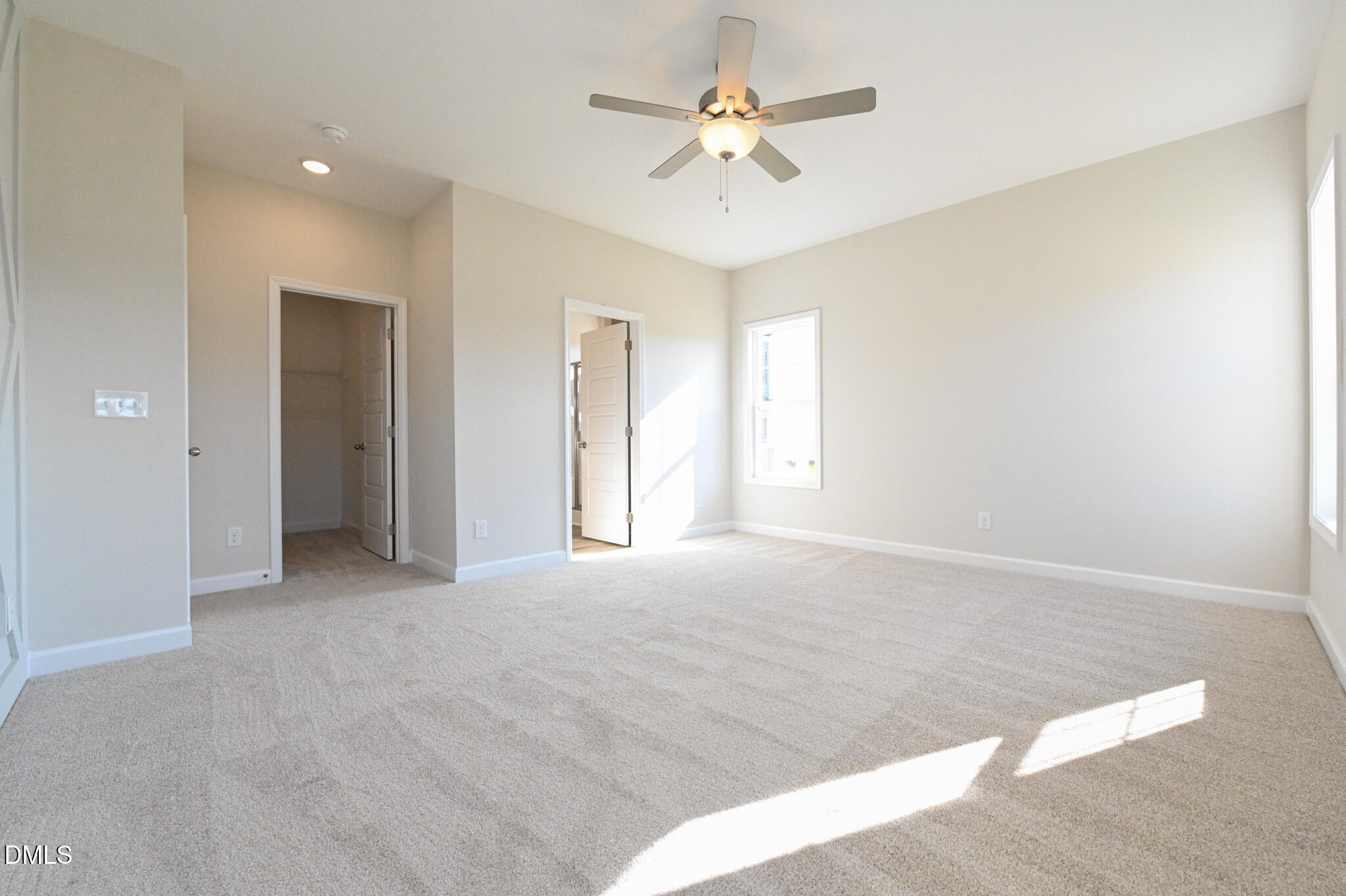 Empty carpeted bedroom with a ceiling fan, two windows, and sunlight across the floor.