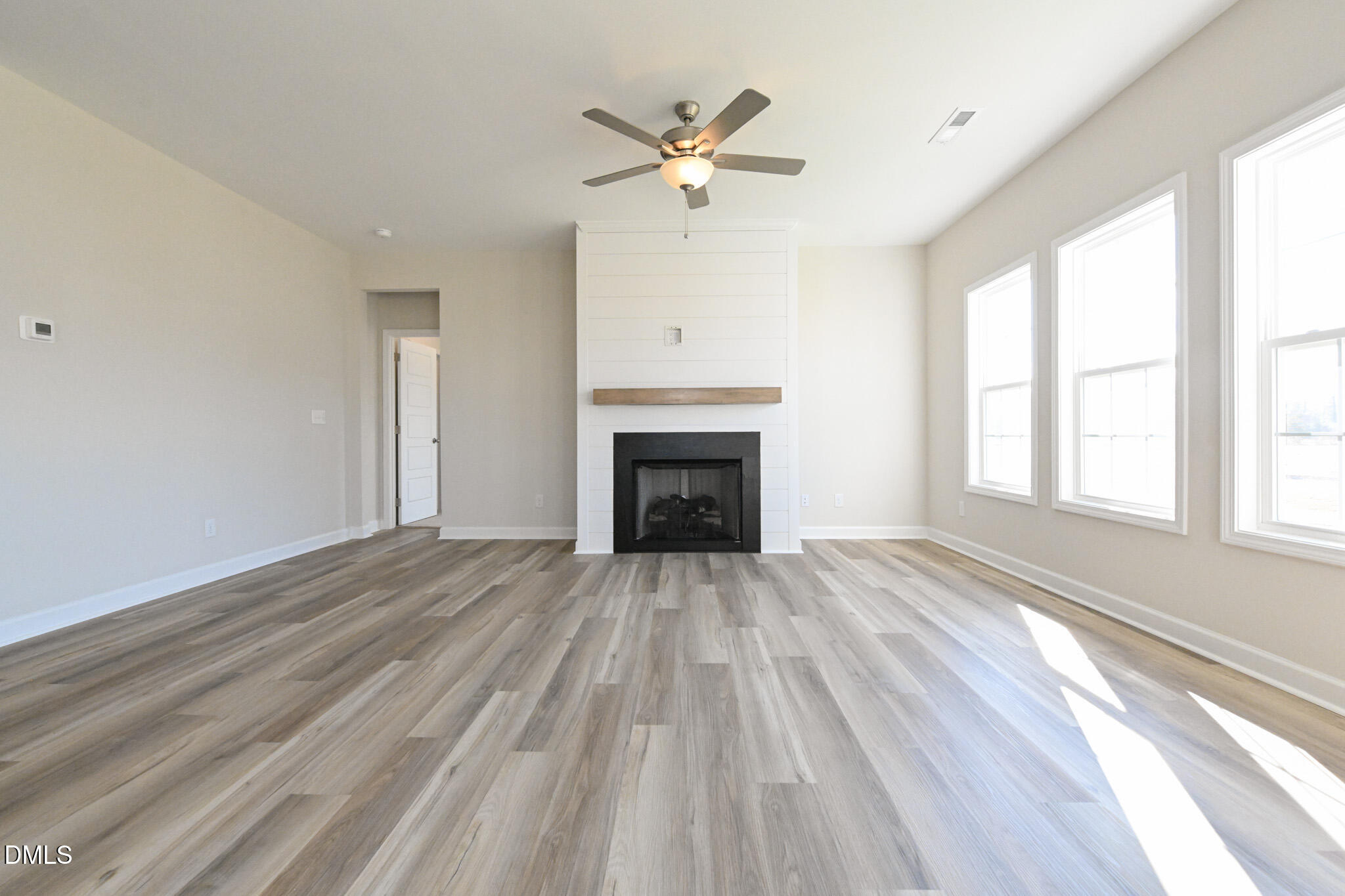 Empty living room with a shiplap fireplace, wood-look floors, ceiling fan, and three tall windows.