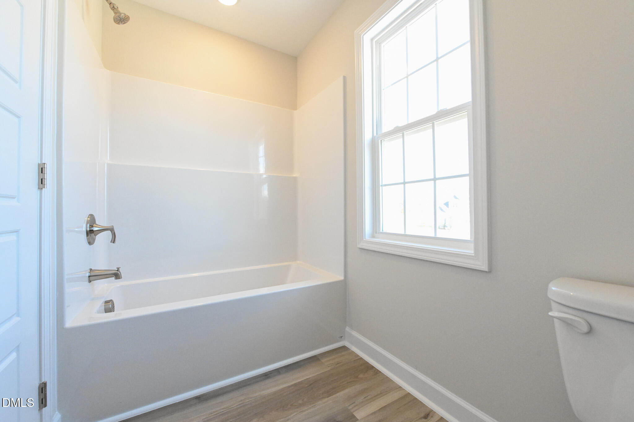 Bathroom with a white tub and shower beside a large window and wood-look flooring.
