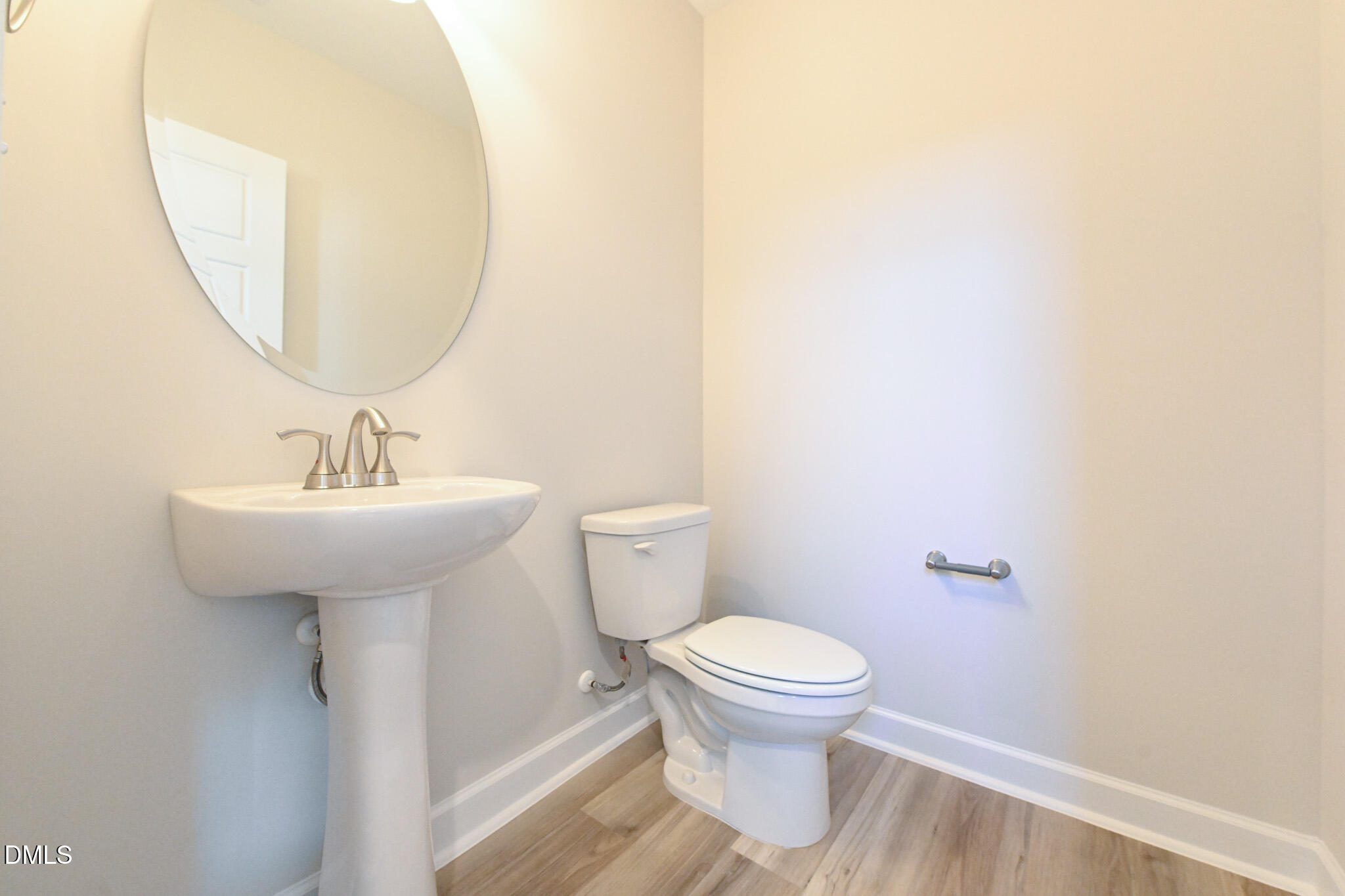 Compact bathroom with a pedestal sink, oval mirror, toilet, and wood-look flooring.