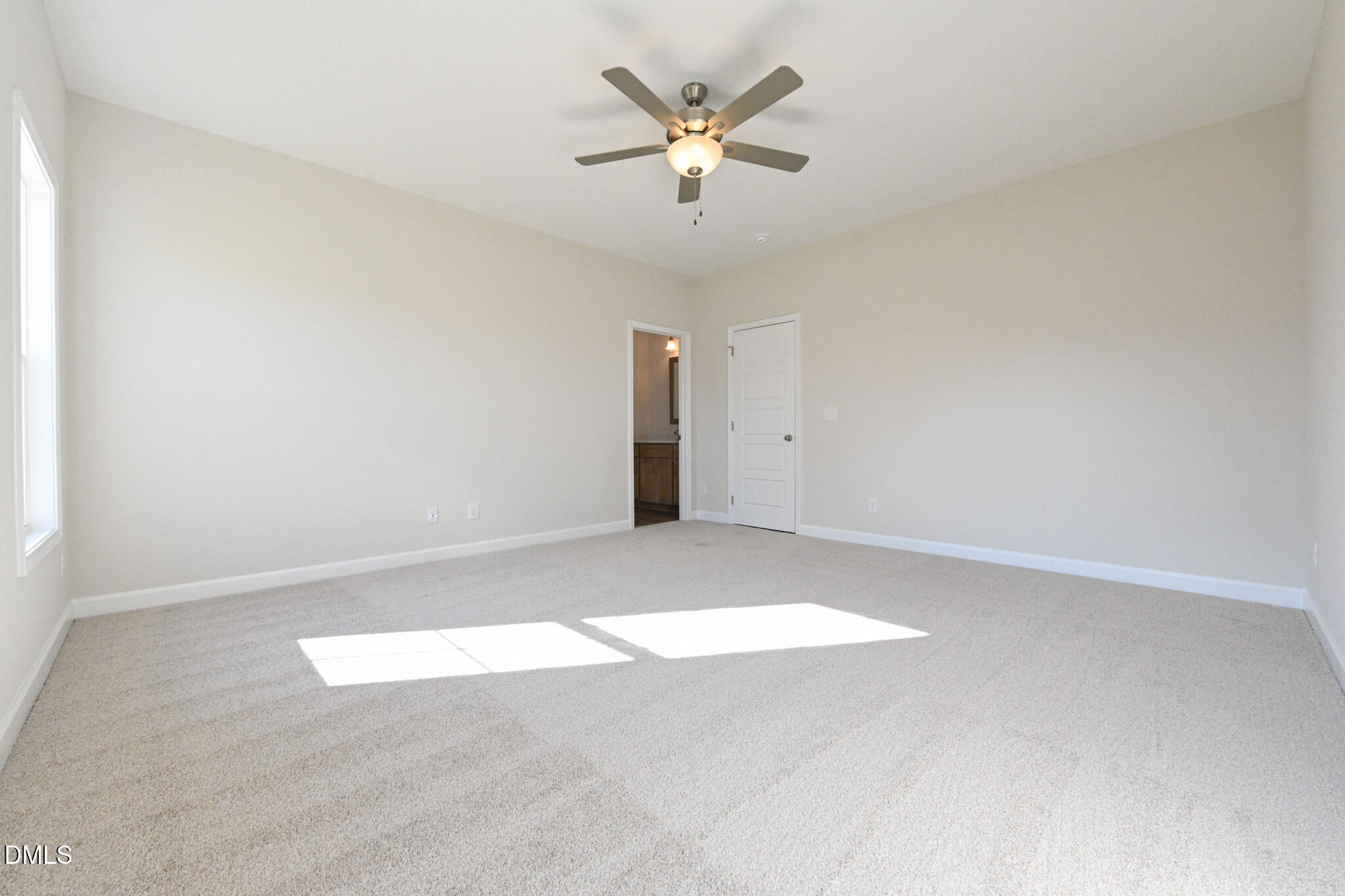 Empty carpeted bedroom with a ceiling fan and bright sunlight streaming through a large window.