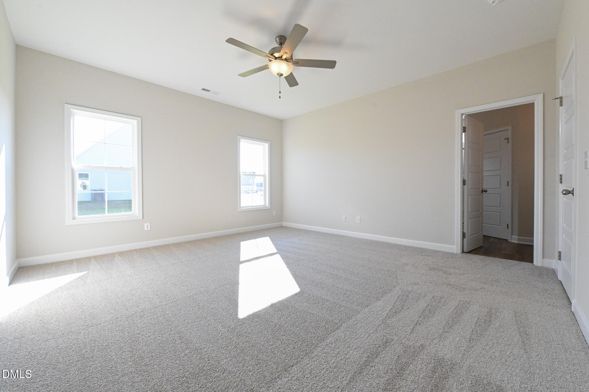 Empty carpeted bedroom with two windows and a ceiling fan, lit by sunlight.