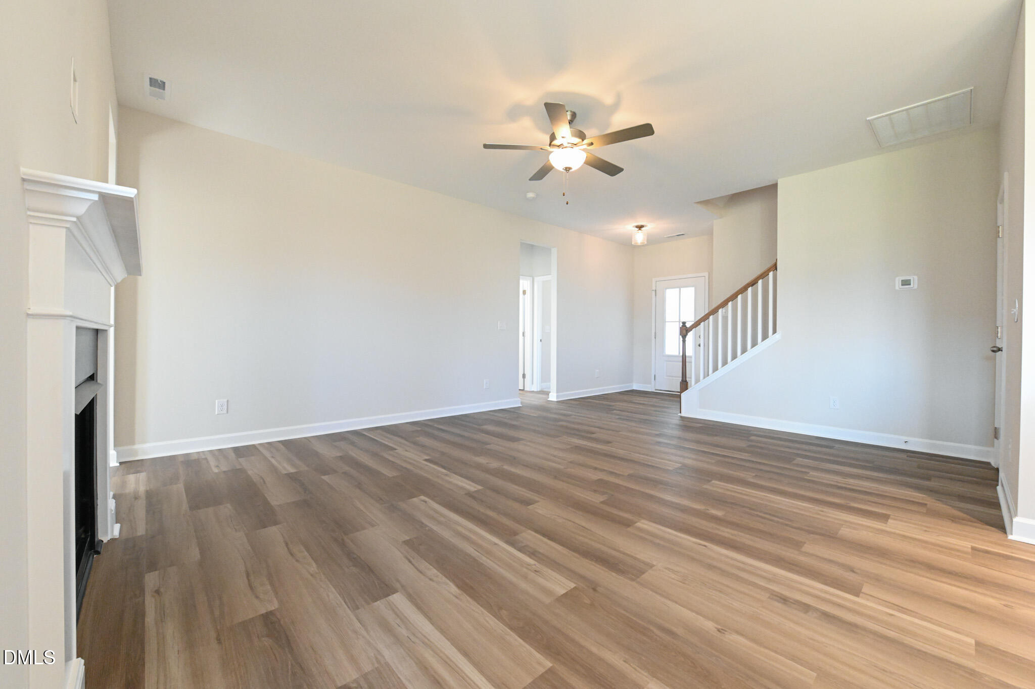 Empty living room with plank flooring, a white fireplace, ceiling fan, and staircase.