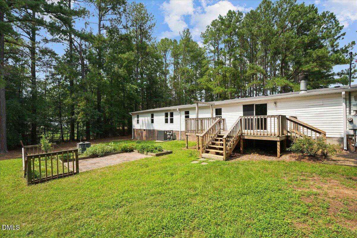 Home’s rear exterior with a raised wooden deck, grassy backyard, patio, and mature trees.