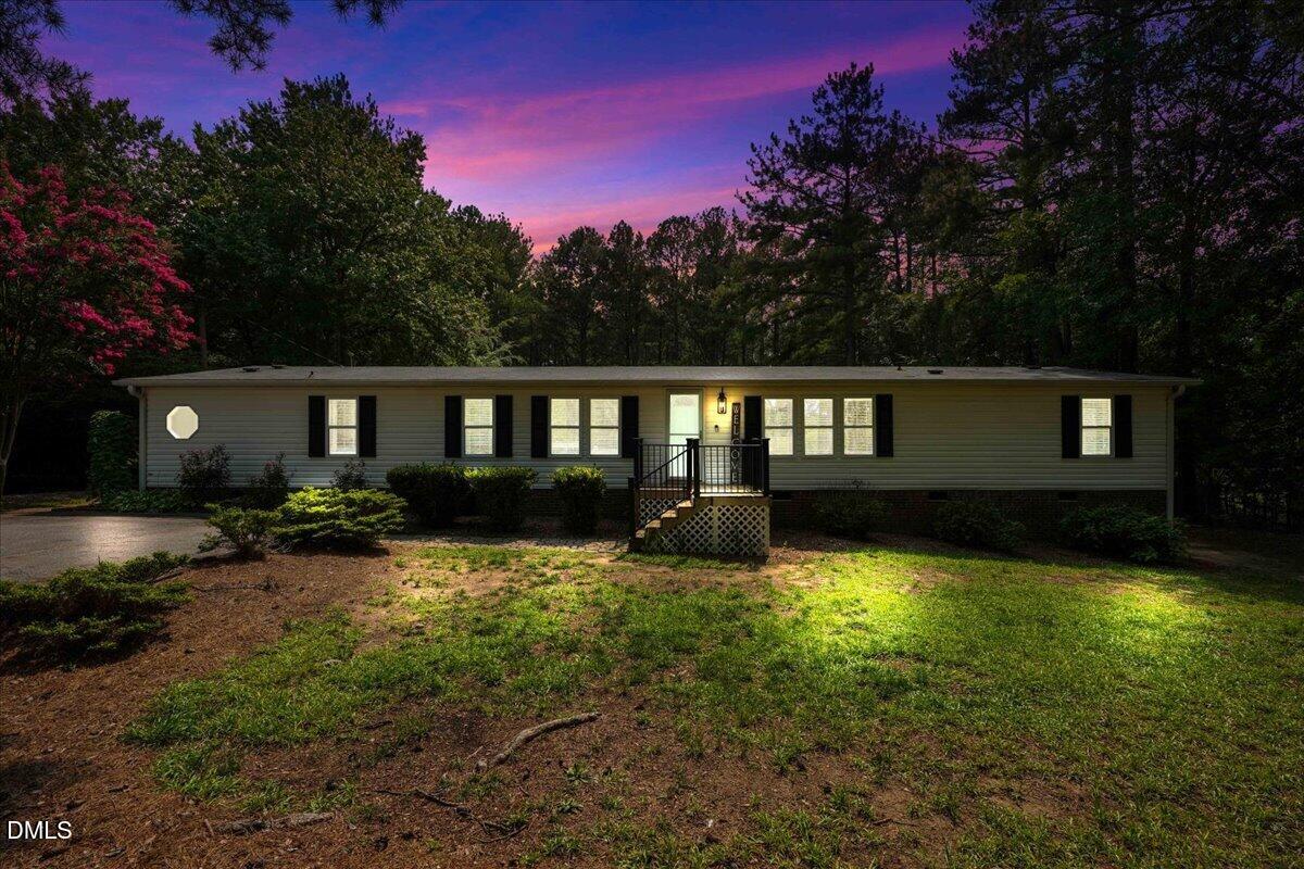 Single-story ranch home at dusk, with illuminated windows, front steps, and wooded surroundings.