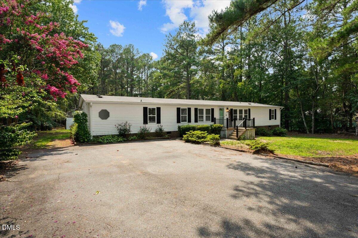 Single-story white home with black shutters, a small porch, and a tree-lined driveway.