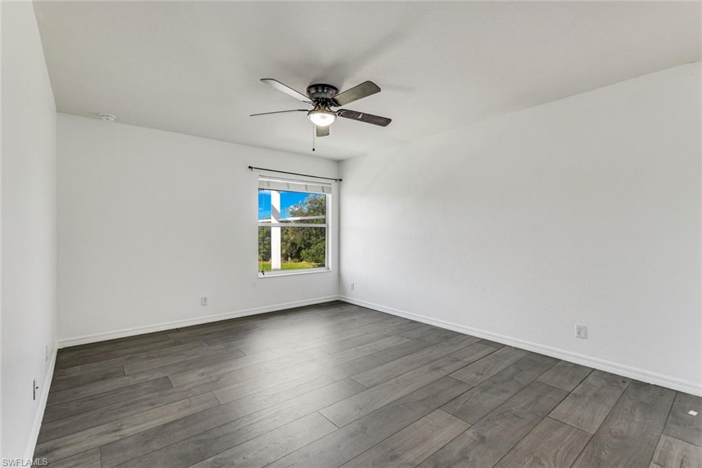 Empty bedroom with gray plank flooring, a ceiling fan, and a window overlooking greenery.