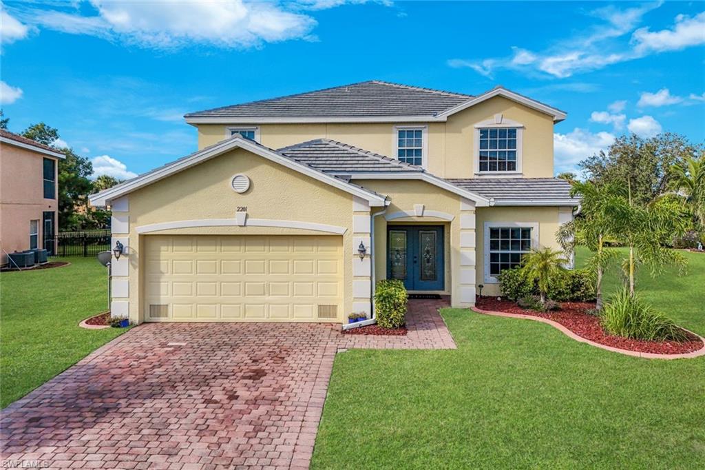 Two-story beige home with a double garage, brick paver driveway, and landscaped front lawn.