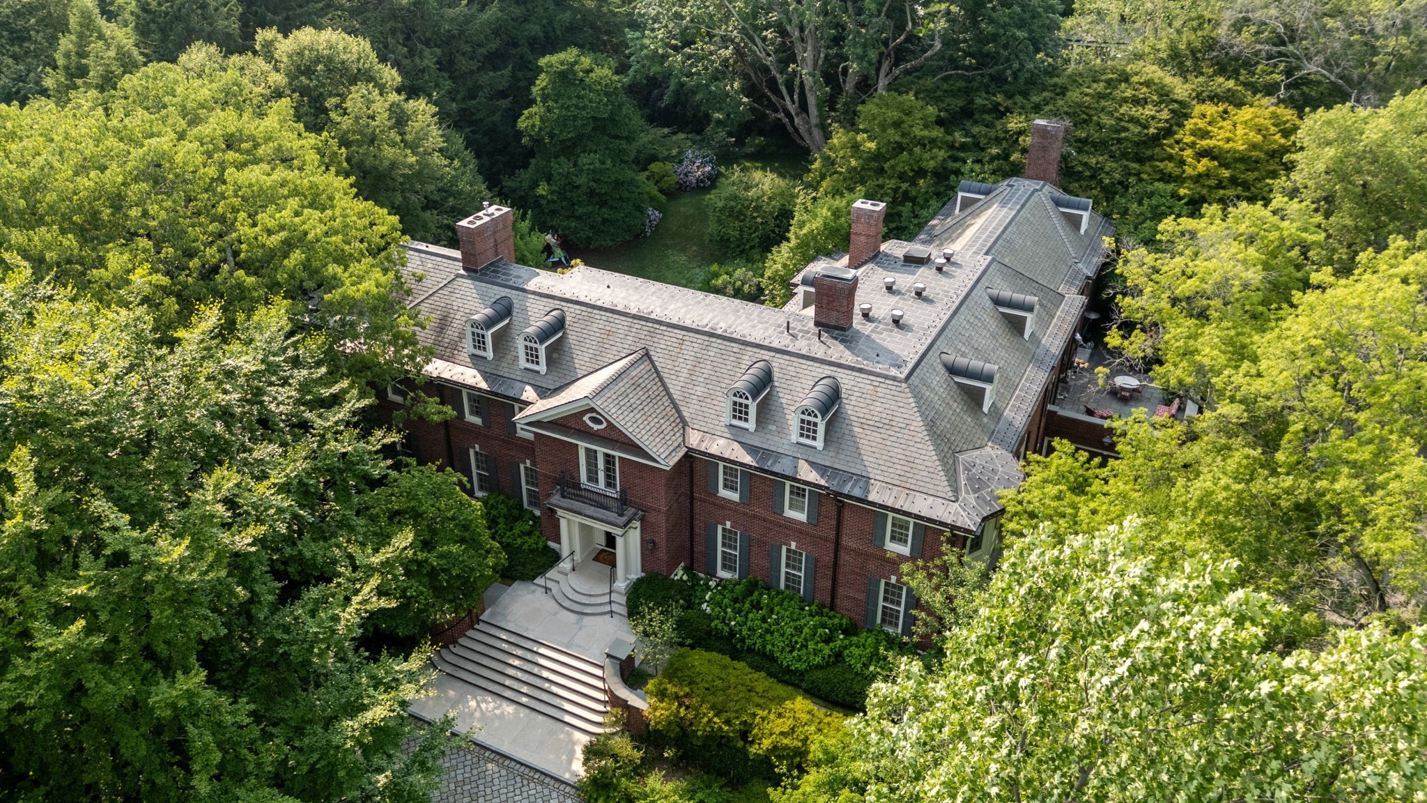 Aerial view of a red-brick home with a slate roof, surrounded by mature trees and landscaped grounds.