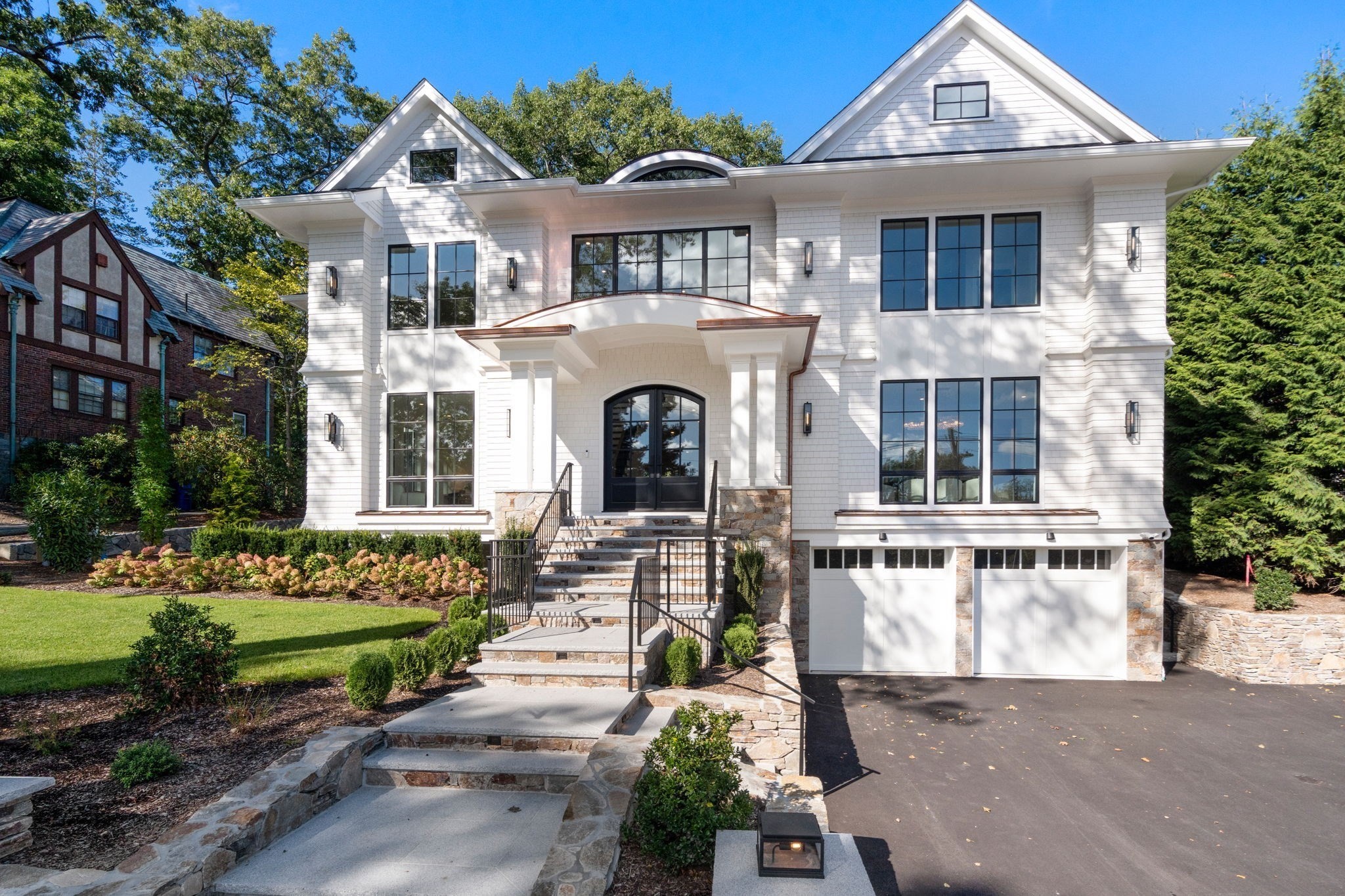 White shingle-style house with black-framed windows, stone entry steps, twin garages, and landscaped front garden.