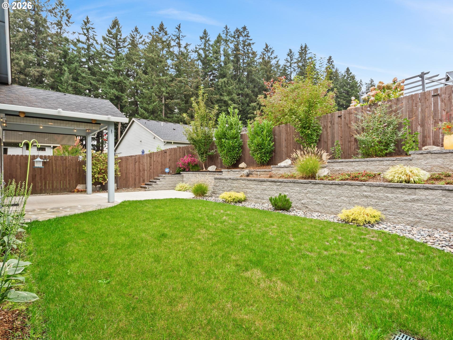 Backyard lawn bordered by tiered stone garden beds, a tall wood fence, and a covered patio.