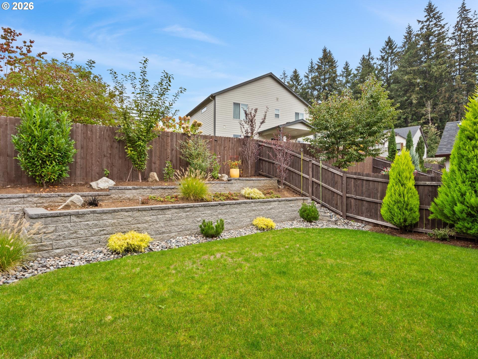 Backyard lawn with tiered stone retaining walls, landscaped beds, and a tall wood fence.