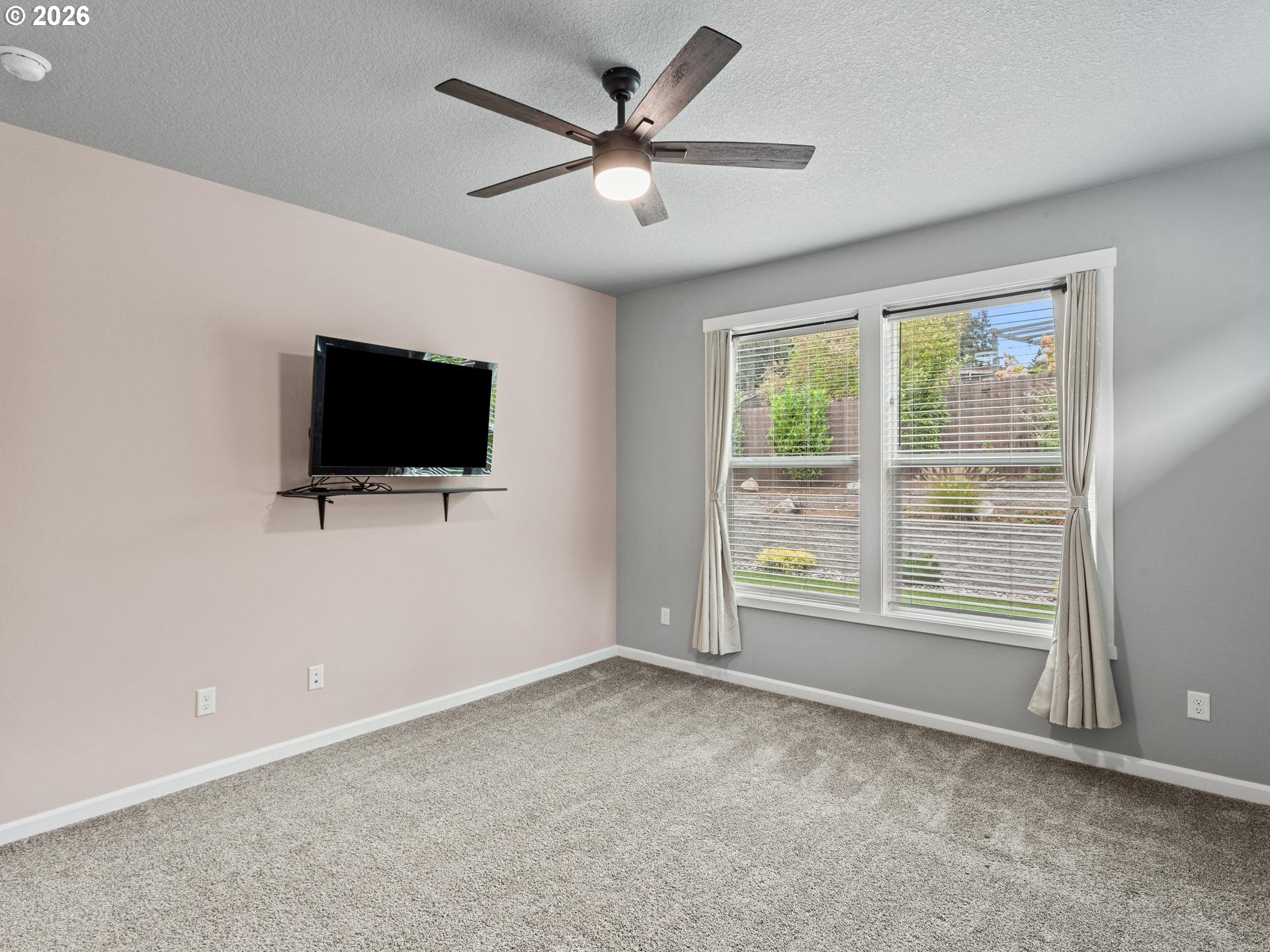 Empty carpeted bedroom with a wall-mounted TV, ceiling fan, and large windows overlooking a garden.
