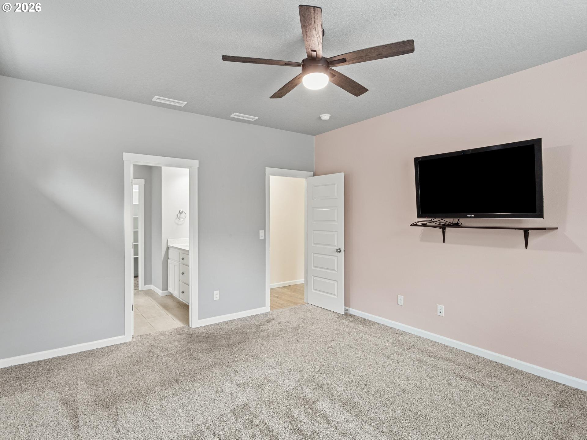 Empty carpeted bedroom with a ceiling fan, wall-mounted television, and adjoining bathroom doorway.