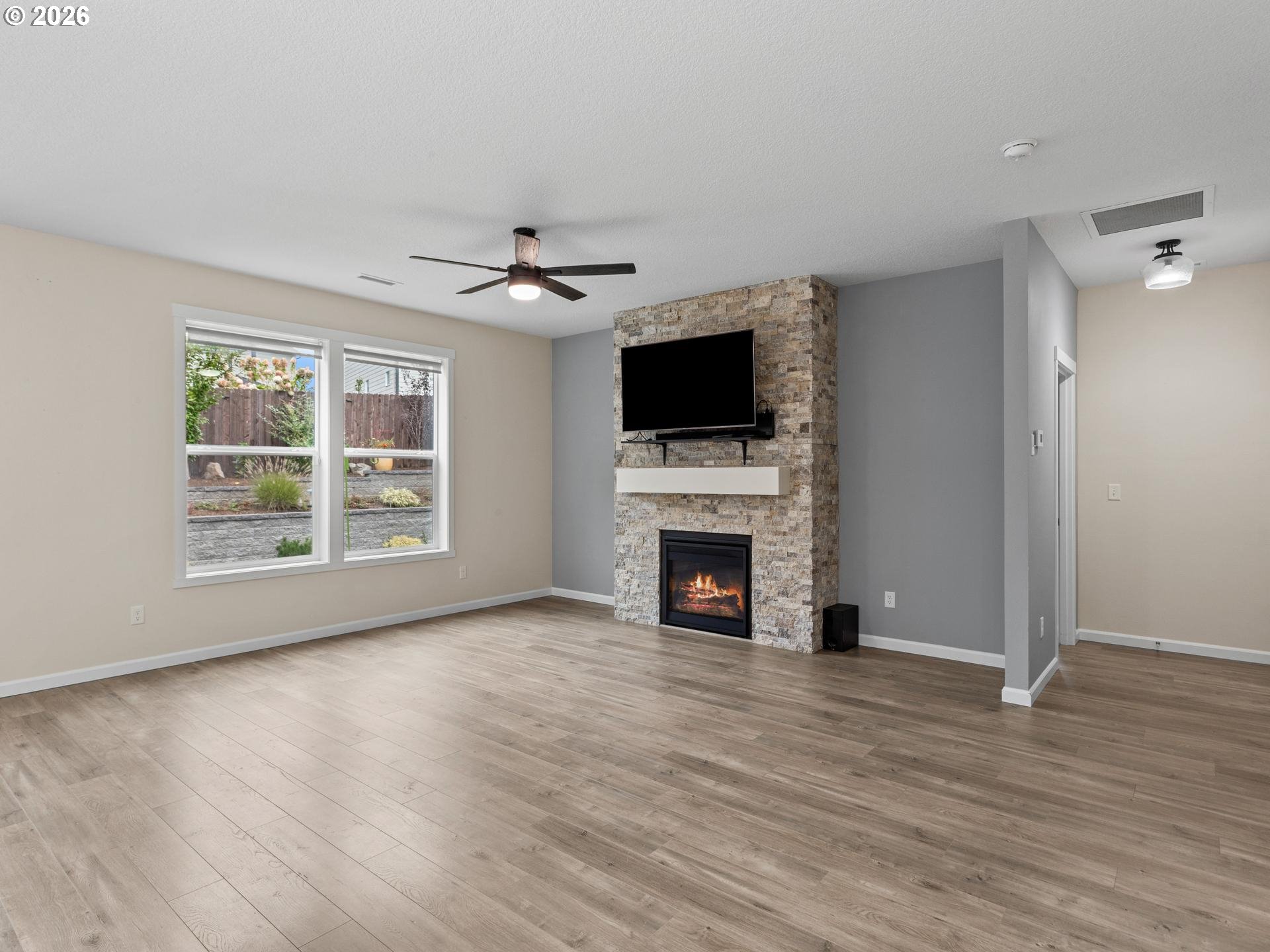 Empty living room with a stone fireplace, mounted TV, and light wood-look floors.