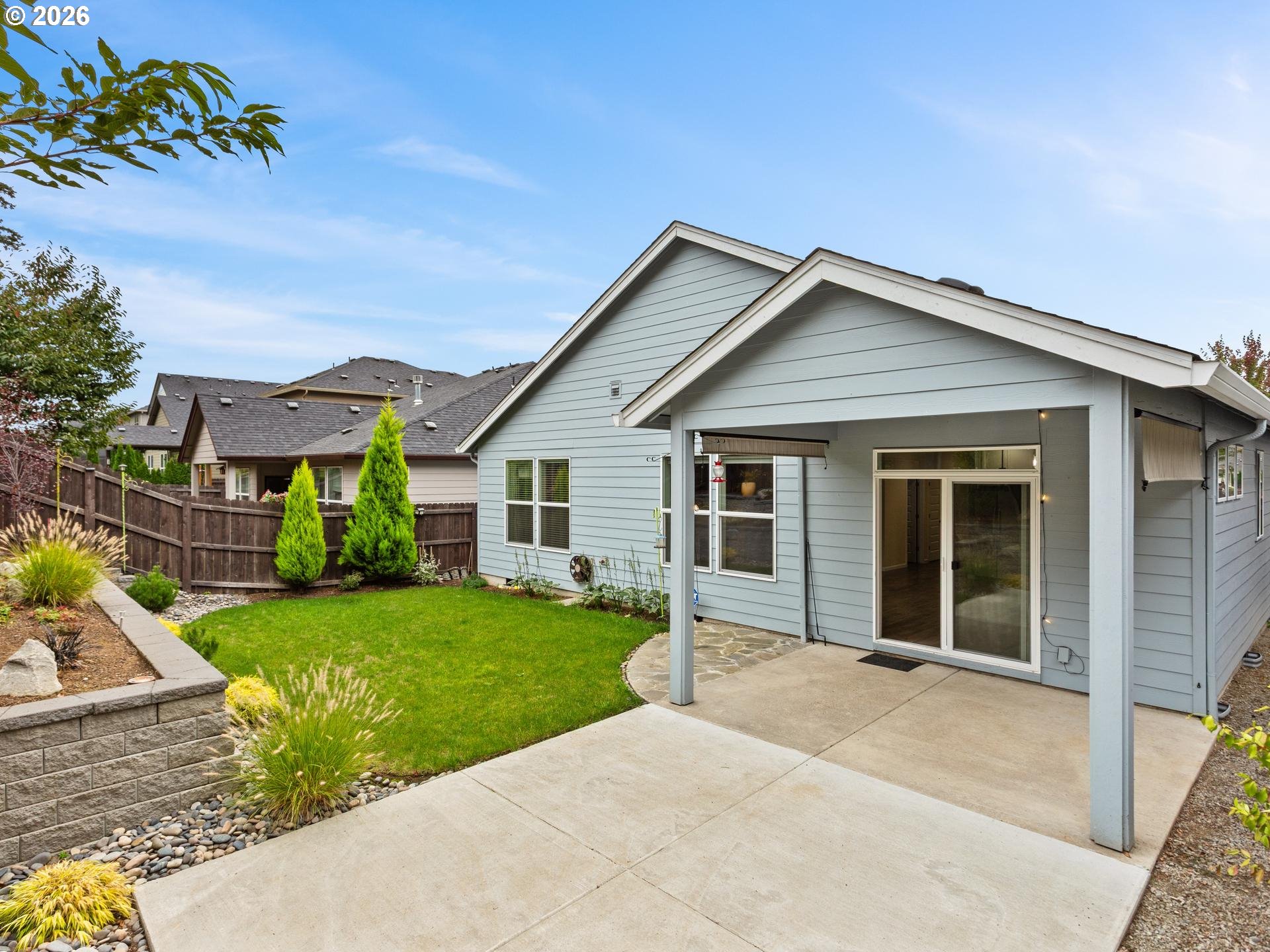 Light blue home’s rear patio and fenced backyard with lawn, stone edging, and covered terrace.