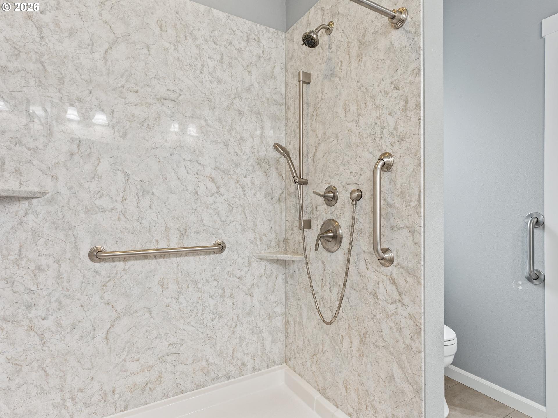 Accessible shower with marble-look walls, grab bars, handheld showerhead, and toilet partly visible.