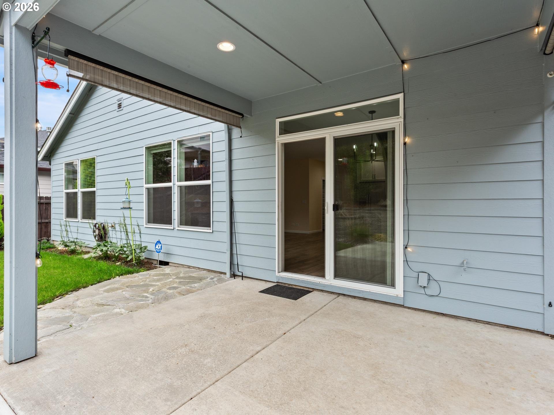 Covered concrete patio with a sliding glass door, overhead lights, and stone walkway beside a blue-sided home.